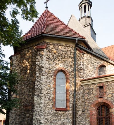 Zdjęcie nr 36: Kościół murowany, orientowany, jednonawowy, trójprzęsłowy, z transeptem i jednoprzęsłowym, węższym i niższym prezbiterium zamkniętym trójbocznie. Między nawą a transeptem po obu stronach znajdują się kruchty, do prezbiterium od strony północnej dostawiona kruchta złożona z dwóch lokalności. Ściany są rozczłonkowane filarami, tworzącymi płytkie arkadowe wnęki. Między nawą a prezbiterium arkada tęczowa. Sklepienie krzyżowe na gurtach spływających na filary. Prezbiterium oświetlają wąskie, prostokątne okna zamknięte ostrołukowo, w nawie i w transepcie znajdują się prostokątne okna prostokątne zamknięte półkoliście, w ścianie frontowej w dolnej części okna prostokątne zamknięte półkoliście, a w górnej podwójne okna prostokątne. Fasada ujęta dwiema narożnymi szkarpami, zwieńczona trójkątnie. Do niej dostawiona kruchta z dwuspadowym daszkiem i wejściem w kształcie prostokąta zamkniętego półkoliście. W kamiennej fasadzie znajdują się dołem okna prostokątne, zamknięte półkoliście, a górą  prostokątne, biforialne okna z nadprożem łukowatym wykonanym z cegły. Szczyt ceglany, podzielony na trzy strefy kamiennym wątkiem, z pięcioma wnękami prostokątnymi, zamkniętymi półkoliście, z wąskimi otworami w dolnej strefie i z owalnym oknem w drugiej. Elewacje boczne nawy ujęte skarpami. Dachy dwuspadowe, kryte dachówką, z wieżyczką na sygnaturkę.         