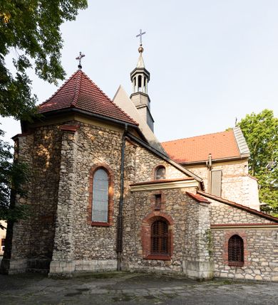 Zdjęcie nr 33: Kościół murowany, orientowany, jednonawowy, trójprzęsłowy, z transeptem i jednoprzęsłowym, węższym i niższym prezbiterium zamkniętym trójbocznie. Między nawą a transeptem po obu stronach znajdują się kruchty, do prezbiterium od strony północnej dostawiona kruchta złożona z dwóch lokalności. Ściany są rozczłonkowane filarami, tworzącymi płytkie arkadowe wnęki. Między nawą a prezbiterium arkada tęczowa. Sklepienie krzyżowe na gurtach spływających na filary. Prezbiterium oświetlają wąskie, prostokątne okna zamknięte ostrołukowo, w nawie i w transepcie znajdują się prostokątne okna prostokątne zamknięte półkoliście, w ścianie frontowej w dolnej części okna prostokątne zamknięte półkoliście, a w górnej podwójne okna prostokątne. Fasada ujęta dwiema narożnymi szkarpami, zwieńczona trójkątnie. Do niej dostawiona kruchta z dwuspadowym daszkiem i wejściem w kształcie prostokąta zamkniętego półkoliście. W kamiennej fasadzie znajdują się dołem okna prostokątne, zamknięte półkoliście, a górą  prostokątne, biforialne okna z nadprożem łukowatym wykonanym z cegły. Szczyt ceglany, podzielony na trzy strefy kamiennym wątkiem, z pięcioma wnękami prostokątnymi, zamkniętymi półkoliście, z wąskimi otworami w dolnej strefie i z owalnym oknem w drugiej. Elewacje boczne nawy ujęte skarpami. Dachy dwuspadowe, kryte dachówką, z wieżyczką na sygnaturkę.         