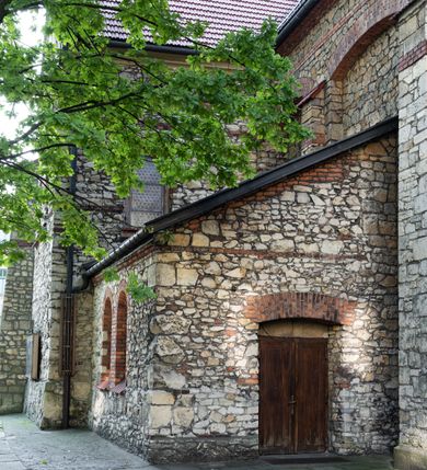 Zdjęcie nr 39: Kościół murowany, orientowany, jednonawowy, trójprzęsłowy, z transeptem i jednoprzęsłowym, węższym i niższym prezbiterium zamkniętym trójbocznie. Między nawą a transeptem po obu stronach znajdują się kruchty, do prezbiterium od strony północnej dostawiona kruchta złożona z dwóch lokalności. Ściany są rozczłonkowane filarami, tworzącymi płytkie arkadowe wnęki. Między nawą a prezbiterium arkada tęczowa. Sklepienie krzyżowe na gurtach spływających na filary. Prezbiterium oświetlają wąskie, prostokątne okna zamknięte ostrołukowo, w nawie i w transepcie znajdują się prostokątne okna prostokątne zamknięte półkoliście, w ścianie frontowej w dolnej części okna prostokątne zamknięte półkoliście, a w górnej podwójne okna prostokątne. Fasada ujęta dwiema narożnymi szkarpami, zwieńczona trójkątnie. Do niej dostawiona kruchta z dwuspadowym daszkiem i wejściem w kształcie prostokąta zamkniętego półkoliście. W kamiennej fasadzie znajdują się dołem okna prostokątne, zamknięte półkoliście, a górą  prostokątne, biforialne okna z nadprożem łukowatym wykonanym z cegły. Szczyt ceglany, podzielony na trzy strefy kamiennym wątkiem, z pięcioma wnękami prostokątnymi, zamkniętymi półkoliście, z wąskimi otworami w dolnej strefie i z owalnym oknem w drugiej. Elewacje boczne nawy ujęte skarpami. Dachy dwuspadowe, kryte dachówką, z wieżyczką na sygnaturkę.         