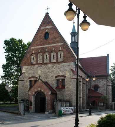 Zdjęcie nr 13: Kościół murowany, orientowany, jednonawowy, trójprzęsłowy, z transeptem i jednoprzęsłowym, węższym i niższym prezbiterium zamkniętym trójbocznie. Między nawą a transeptem po obu stronach znajdują się kruchty, do prezbiterium od strony północnej dostawiona kruchta złożona z dwóch lokalności. Ściany są rozczłonkowane filarami, tworzącymi płytkie arkadowe wnęki. Między nawą a prezbiterium arkada tęczowa. Sklepienie krzyżowe na gurtach spływających na filary. Prezbiterium oświetlają wąskie, prostokątne okna zamknięte ostrołukowo, w nawie i w transepcie znajdują się prostokątne okna prostokątne zamknięte półkoliście, w ścianie frontowej w dolnej części okna prostokątne zamknięte półkoliście, a w górnej podwójne okna prostokątne. Fasada ujęta dwiema narożnymi szkarpami, zwieńczona trójkątnie. Do niej dostawiona kruchta z dwuspadowym daszkiem i wejściem w kształcie prostokąta zamkniętego półkoliście. W kamiennej fasadzie znajdują się dołem okna prostokątne, zamknięte półkoliście, a górą  prostokątne, biforialne okna z nadprożem łukowatym wykonanym z cegły. Szczyt ceglany, podzielony na trzy strefy kamiennym wątkiem, z pięcioma wnękami prostokątnymi, zamkniętymi półkoliście, z wąskimi otworami w dolnej strefie i z owalnym oknem w drugiej. Elewacje boczne nawy ujęte skarpami. Dachy dwuspadowe, kryte dachówką, z wieżyczką na sygnaturkę.         