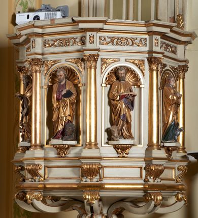 Zdjęcie nr 2: Ambona nadwieszona, przyścienna. Jej kosz jest ośmioboczny, wsparty na szerokich wolutach z plintą w kształcie winnego grona. Ścianki kosza podzielone kompozytowymi półkolumnami wspartymi na konsolkach z liściastymi wolutami. Między kolumienkami nisze konchowe z muszlami, wewnątrz których postacie ewangelistów, od lewej Mateusza, Marka, Łukasza i Jana. Figury ewangelistów ujęte całopostaciowo w pozycji stojącej, ubrane w długie tuniki i płaszcze. W dłoniach podtrzymują otwarte księgi, a u stóp widnieją ich symbole. Kosz wieńczy belkowanie wyłamujące się ponad kolumienkami, z fryzem zdobionym wiciami akantu. Zaplecek z dwiema płycinami zamkniętymi łukiem półkolistym. Ponad nimi pole w kształcie leżącego prostokąta, wewnątrz którego krzyż. Zaplecek flankowany dwiema listwami ujętymi ulistnionymi konsolami. Pośrodku dwie płyciny w kształcie wąskich prostokątów zakończonych półkoliście. W górnej części prostokątne pole w złoconym profilu z listkami wypełniającymi górne naroża, z przerwanym, wolutowym przyczółkiem i krzyżem o ramionach zakończonych trójlistnie. Baldachim na rzucie ośmioboku, z podwieszonym lambrekinem z chwostami i z ażurową koronką w kształcie wici roślinnej. Na baldachimie ośmioboczny cokół uskokowy, z płycinami w kształcie stojących prostokątów zamkniętych łukiem półkolistym nadwieszonym. Na cokole pełnoplastyczna figura stojącego Jana Chrzciciela, ubranego w wielbłądzią skórę i złocony płaszcz przerzucony przez lewe ramię. Jego twarz jest pociągła, okolona zarostem, włosy sięgające uszu. Prawą rękę unosi w geście błogosławieństwa, a w lewej trzyma krzyż z banderolą. W podniebiu gołębica Ducha Świętego na tle glorii promienistej. Ambona polichromowana na szaro, detale złocone. 
