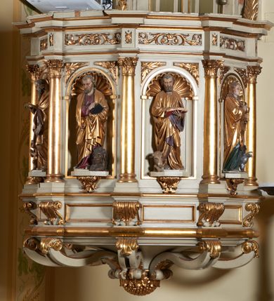 Zdjęcie nr 1: Ambona nadwieszona, przyścienna. Jej kosz jest ośmioboczny, wsparty na szerokich wolutach z plintą w kształcie winnego grona. Ścianki kosza podzielone kompozytowymi półkolumnami wspartymi na konsolkach z liściastymi wolutami. Między kolumienkami nisze konchowe z muszlami, wewnątrz których postacie ewangelistów, od lewej Mateusza, Marka, Łukasza i Jana. Figury ewangelistów ujęte całopostaciowo w pozycji stojącej, ubrane w długie tuniki i płaszcze. W dłoniach podtrzymują otwarte księgi, a u stóp widnieją ich symbole. Kosz wieńczy belkowanie wyłamujące się ponad kolumienkami, z fryzem zdobionym wiciami akantu. Zaplecek z dwiema płycinami zamkniętymi łukiem półkolistym. Ponad nimi pole w kształcie leżącego prostokąta, wewnątrz którego krzyż. Zaplecek flankowany dwiema listwami ujętymi ulistnionymi konsolami. Pośrodku dwie płyciny w kształcie wąskich prostokątów zakończonych półkoliście. W górnej części prostokątne pole w złoconym profilu z listkami wypełniającymi górne naroża, z przerwanym, wolutowym przyczółkiem i krzyżem o ramionach zakończonych trójlistnie. Baldachim na rzucie ośmioboku, z podwieszonym lambrekinem z chwostami i z ażurową koronką w kształcie wici roślinnej. Na baldachimie ośmioboczny cokół uskokowy, z płycinami w kształcie stojących prostokątów zamkniętych łukiem półkolistym nadwieszonym. Na cokole pełnoplastyczna figura stojącego Jana Chrzciciela, ubranego w wielbłądzią skórę i złocony płaszcz przerzucony przez lewe ramię. Jego twarz jest pociągła, okolona zarostem, włosy sięgające uszu. Prawą rękę unosi w geście błogosławieństwa, a w lewej trzyma krzyż z banderolą. W podniebiu gołębica Ducha Świętego na tle glorii promienistej. Ambona polichromowana na szaro, detale złocone. 
