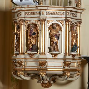 Zdjęcie nr 1: Ambona nadwieszona, przyścienna. Jej kosz jest ośmioboczny, wsparty na szerokich wolutach z plintą w kształcie winnego grona. Ścianki kosza podzielone kompozytowymi półkolumnami wspartymi na konsolkach z liściastymi wolutami. Między kolumienkami nisze konchowe z muszlami, wewnątrz których postacie ewangelistów, od lewej Mateusza, Marka, Łukasza i Jana. Figury ewangelistów ujęte całopostaciowo w pozycji stojącej, ubrane w długie tuniki i płaszcze. W dłoniach podtrzymują otwarte księgi, a u stóp widnieją ich symbole. Kosz wieńczy belkowanie wyłamujące się ponad kolumienkami, z fryzem zdobionym wiciami akantu. Zaplecek z dwiema płycinami zamkniętymi łukiem półkolistym. Ponad nimi pole w kształcie leżącego prostokąta, wewnątrz którego krzyż. Zaplecek flankowany dwiema listwami ujętymi ulistnionymi konsolami. Pośrodku dwie płyciny w kształcie wąskich prostokątów zakończonych półkoliście. W górnej części prostokątne pole w złoconym profilu z listkami wypełniającymi górne naroża, z przerwanym, wolutowym przyczółkiem i krzyżem o ramionach zakończonych trójlistnie. Baldachim na rzucie ośmioboku, z podwieszonym lambrekinem z chwostami i z ażurową koronką w kształcie wici roślinnej. Na baldachimie ośmioboczny cokół uskokowy, z płycinami w kształcie stojących prostokątów zamkniętych łukiem półkolistym nadwieszonym. Na cokole pełnoplastyczna figura stojącego Jana Chrzciciela, ubranego w wielbłądzią skórę i złocony płaszcz przerzucony przez lewe ramię. Jego twarz jest pociągła, okolona zarostem, włosy sięgające uszu. Prawą rękę unosi w geście błogosławieństwa, a w lewej trzyma krzyż z banderolą. W podniebiu gołębica Ducha Świętego na tle glorii promienistej. Ambona polichromowana na szaro, detale złocone. 
