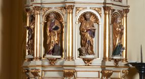 Zdjęcie nr 1: Ambona nadwieszona, przyścienna. Jej kosz jest ośmioboczny, wsparty na szerokich wolutach z plintą w kształcie winnego grona. Ścianki kosza podzielone kompozytowymi półkolumnami wspartymi na konsolkach z liściastymi wolutami. Między kolumienkami nisze konchowe z muszlami, wewnątrz których postacie ewangelistów, od lewej Mateusza, Marka, Łukasza i Jana. Figury ewangelistów ujęte całopostaciowo w pozycji stojącej, ubrane w długie tuniki i płaszcze. W dłoniach podtrzymują otwarte księgi, a u stóp widnieją ich symbole. Kosz wieńczy belkowanie wyłamujące się ponad kolumienkami, z fryzem zdobionym wiciami akantu. Zaplecek z dwiema płycinami zamkniętymi łukiem półkolistym. Ponad nimi pole w kształcie leżącego prostokąta, wewnątrz którego krzyż. Zaplecek flankowany dwiema listwami ujętymi ulistnionymi konsolami. Pośrodku dwie płyciny w kształcie wąskich prostokątów zakończonych półkoliście. W górnej części prostokątne pole w złoconym profilu z listkami wypełniającymi górne naroża, z przerwanym, wolutowym przyczółkiem i krzyżem o ramionach zakończonych trójlistnie. Baldachim na rzucie ośmioboku, z podwieszonym lambrekinem z chwostami i z ażurową koronką w kształcie wici roślinnej. Na baldachimie ośmioboczny cokół uskokowy, z płycinami w kształcie stojących prostokątów zamkniętych łukiem półkolistym nadwieszonym. Na cokole pełnoplastyczna figura stojącego Jana Chrzciciela, ubranego w wielbłądzią skórę i złocony płaszcz przerzucony przez lewe ramię. Jego twarz jest pociągła, okolona zarostem, włosy sięgające uszu. Prawą rękę unosi w geście błogosławieństwa, a w lewej trzyma krzyż z banderolą. W podniebiu gołębica Ducha Świętego na tle glorii promienistej. Ambona polichromowana na szaro, detale złocone. 
