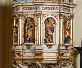 Zdjęcie nr 1: Ambona nadwieszona, przyścienna. Jej kosz jest ośmioboczny, wsparty na szerokich wolutach z plintą w kształcie winnego grona. Ścianki kosza podzielone kompozytowymi półkolumnami wspartymi na konsolkach z liściastymi wolutami. Między kolumienkami nisze konchowe z muszlami, wewnątrz których postacie ewangelistów, od lewej Mateusza, Marka, Łukasza i Jana. Figury ewangelistów ujęte całopostaciowo w pozycji stojącej, ubrane w długie tuniki i płaszcze. W dłoniach podtrzymują otwarte księgi, a u stóp widnieją ich symbole. Kosz wieńczy belkowanie wyłamujące się ponad kolumienkami, z fryzem zdobionym wiciami akantu. Zaplecek z dwiema płycinami zamkniętymi łukiem półkolistym. Ponad nimi pole w kształcie leżącego prostokąta, wewnątrz którego krzyż. Zaplecek flankowany dwiema listwami ujętymi ulistnionymi konsolami. Pośrodku dwie płyciny w kształcie wąskich prostokątów zakończonych półkoliście. W górnej części prostokątne pole w złoconym profilu z listkami wypełniającymi górne naroża, z przerwanym, wolutowym przyczółkiem i krzyżem o ramionach zakończonych trójlistnie. Baldachim na rzucie ośmioboku, z podwieszonym lambrekinem z chwostami i z ażurową koronką w kształcie wici roślinnej. Na baldachimie ośmioboczny cokół uskokowy, z płycinami w kształcie stojących prostokątów zamkniętych łukiem półkolistym nadwieszonym. Na cokole pełnoplastyczna figura stojącego Jana Chrzciciela, ubranego w wielbłądzią skórę i złocony płaszcz przerzucony przez lewe ramię. Jego twarz jest pociągła, okolona zarostem, włosy sięgające uszu. Prawą rękę unosi w geście błogosławieństwa, a w lewej trzyma krzyż z banderolą. W podniebiu gołębica Ducha Świętego na tle glorii promienistej. Ambona polichromowana na szaro, detale złocone. 
