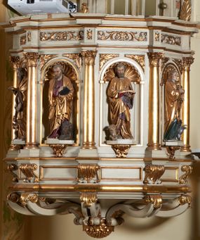 Zdjęcie nr 1: Ambona nadwieszona, przyścienna. Jej kosz jest ośmioboczny, wsparty na szerokich wolutach z plintą w kształcie winnego grona. Ścianki kosza podzielone kompozytowymi półkolumnami wspartymi na konsolkach z liściastymi wolutami. Między kolumienkami nisze konchowe z muszlami, wewnątrz których postacie ewangelistów, od lewej Mateusza, Marka, Łukasza i Jana. Figury ewangelistów ujęte całopostaciowo w pozycji stojącej, ubrane w długie tuniki i płaszcze. W dłoniach podtrzymują otwarte księgi, a u stóp widnieją ich symbole. Kosz wieńczy belkowanie wyłamujące się ponad kolumienkami, z fryzem zdobionym wiciami akantu. Zaplecek z dwiema płycinami zamkniętymi łukiem półkolistym. Ponad nimi pole w kształcie leżącego prostokąta, wewnątrz którego krzyż. Zaplecek flankowany dwiema listwami ujętymi ulistnionymi konsolami. Pośrodku dwie płyciny w kształcie wąskich prostokątów zakończonych półkoliście. W górnej części prostokątne pole w złoconym profilu z listkami wypełniającymi górne naroża, z przerwanym, wolutowym przyczółkiem i krzyżem o ramionach zakończonych trójlistnie. Baldachim na rzucie ośmioboku, z podwieszonym lambrekinem z chwostami i z ażurową koronką w kształcie wici roślinnej. Na baldachimie ośmioboczny cokół uskokowy, z płycinami w kształcie stojących prostokątów zamkniętych łukiem półkolistym nadwieszonym. Na cokole pełnoplastyczna figura stojącego Jana Chrzciciela, ubranego w wielbłądzią skórę i złocony płaszcz przerzucony przez lewe ramię. Jego twarz jest pociągła, okolona zarostem, włosy sięgające uszu. Prawą rękę unosi w geście błogosławieństwa, a w lewej trzyma krzyż z banderolą. W podniebiu gołębica Ducha Świętego na tle glorii promienistej. Ambona polichromowana na szaro, detale złocone. 
