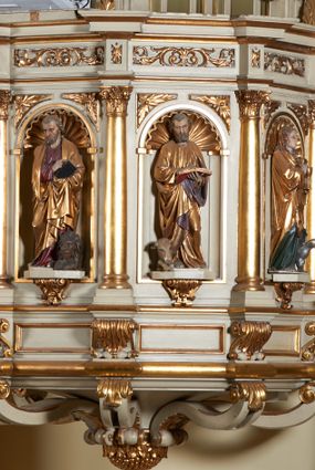 Zdjęcie nr 1: Ambona nadwieszona, przyścienna. Jej kosz jest ośmioboczny, wsparty na szerokich wolutach z plintą w kształcie winnego grona. Ścianki kosza podzielone kompozytowymi półkolumnami wspartymi na konsolkach z liściastymi wolutami. Między kolumienkami nisze konchowe z muszlami, wewnątrz których postacie ewangelistów, od lewej Mateusza, Marka, Łukasza i Jana. Figury ewangelistów ujęte całopostaciowo w pozycji stojącej, ubrane w długie tuniki i płaszcze. W dłoniach podtrzymują otwarte księgi, a u stóp widnieją ich symbole. Kosz wieńczy belkowanie wyłamujące się ponad kolumienkami, z fryzem zdobionym wiciami akantu. Zaplecek z dwiema płycinami zamkniętymi łukiem półkolistym. Ponad nimi pole w kształcie leżącego prostokąta, wewnątrz którego krzyż. Zaplecek flankowany dwiema listwami ujętymi ulistnionymi konsolami. Pośrodku dwie płyciny w kształcie wąskich prostokątów zakończonych półkoliście. W górnej części prostokątne pole w złoconym profilu z listkami wypełniającymi górne naroża, z przerwanym, wolutowym przyczółkiem i krzyżem o ramionach zakończonych trójlistnie. Baldachim na rzucie ośmioboku, z podwieszonym lambrekinem z chwostami i z ażurową koronką w kształcie wici roślinnej. Na baldachimie ośmioboczny cokół uskokowy, z płycinami w kształcie stojących prostokątów zamkniętych łukiem półkolistym nadwieszonym. Na cokole pełnoplastyczna figura stojącego Jana Chrzciciela, ubranego w wielbłądzią skórę i złocony płaszcz przerzucony przez lewe ramię. Jego twarz jest pociągła, okolona zarostem, włosy sięgające uszu. Prawą rękę unosi w geście błogosławieństwa, a w lewej trzyma krzyż z banderolą. W podniebiu gołębica Ducha Świętego na tle glorii promienistej. Ambona polichromowana na szaro, detale złocone. 
