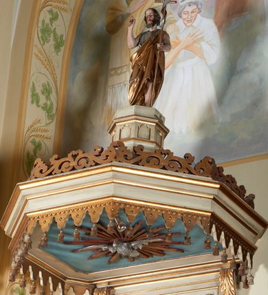 Zdjęcie nr 3: Ambona nadwieszona, przyścienna. Jej kosz jest ośmioboczny, wsparty na szerokich wolutach z plintą w kształcie winnego grona. Ścianki kosza podzielone kompozytowymi półkolumnami wspartymi na konsolkach z liściastymi wolutami. Między kolumienkami nisze konchowe z muszlami, wewnątrz których postacie ewangelistów, od lewej Mateusza, Marka, Łukasza i Jana. Figury ewangelistów ujęte całopostaciowo w pozycji stojącej, ubrane w długie tuniki i płaszcze. W dłoniach podtrzymują otwarte księgi, a u stóp widnieją ich symbole. Kosz wieńczy belkowanie wyłamujące się ponad kolumienkami, z fryzem zdobionym wiciami akantu. Zaplecek z dwiema płycinami zamkniętymi łukiem półkolistym. Ponad nimi pole w kształcie leżącego prostokąta, wewnątrz którego krzyż. Zaplecek flankowany dwiema listwami ujętymi ulistnionymi konsolami. Pośrodku dwie płyciny w kształcie wąskich prostokątów zakończonych półkoliście. W górnej części prostokątne pole w złoconym profilu z listkami wypełniającymi górne naroża, z przerwanym, wolutowym przyczółkiem i krzyżem o ramionach zakończonych trójlistnie. Baldachim na rzucie ośmioboku, z podwieszonym lambrekinem z chwostami i z ażurową koronką w kształcie wici roślinnej. Na baldachimie ośmioboczny cokół uskokowy, z płycinami w kształcie stojących prostokątów zamkniętych łukiem półkolistym nadwieszonym. Na cokole pełnoplastyczna figura stojącego Jana Chrzciciela, ubranego w wielbłądzią skórę i złocony płaszcz przerzucony przez lewe ramię. Jego twarz jest pociągła, okolona zarostem, włosy sięgające uszu. Prawą rękę unosi w geście błogosławieństwa, a w lewej trzyma krzyż z banderolą. W podniebiu gołębica Ducha Świętego na tle glorii promienistej. Ambona polichromowana na szaro, detale złocone. 
