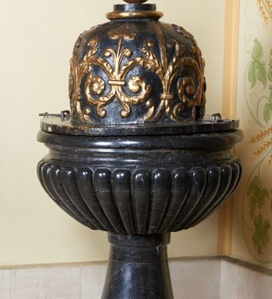 Zdjęcie nr 1: Chrzcielnica osadzona na okrągłej podstawie i prostopadłościennym, spłaszczonym cokole oraz cylindrycznym w przekroju trzonie, dołem lekko wybrzuszonym. Czara półkulista, puklowana, z profilowanym wylewem. Pokrywa drewniana, na okrągłej podstawie, cylindryczna, zaoblona u góry, zwieńczona wałkiem i kulą z krzyżem. Jej powierzchnia jest pokryta biegnącymi wzdłuż, złoconymi czterema taśmami i odchodzącymi od nich wolutowymi, spiętymi ze sobą gałązkami. 