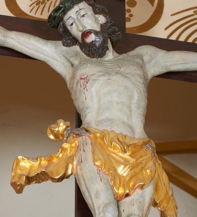 Zdjęcie nr 7: Grupa Ukrzyżowania złożona z ukrzyżowanego Chrystusa w typie christo vivo, stojących postaci Matki Boskiej i św. Jana oraz klęczącej Marii Magdaleny. Figura Chrystusa jest rzeźbą pełnoplastyczną, nieznacznie ściętą z tyłu, pozostałe są ścięte i wydrążone od tyłu. Zbawiciel przybity trzema gwoździami, z niemal prosto rozpostartymi rękami, z głową przechyloną na prawe ramię. Jego twarz jest pociągła, nos długi, oczy otwarte, twarz okolona kręconą, brązową brodą na głowie kręcone, sięgające szyi włosy i korona cierniowa pomalowana na zielono. Ręce z wyłamanymi barkami, z uwidocznionymi ścięgnami, klatka piersiowa wybrzuszona, z widocznymi żebrami i mostkiem, nogi smukłe z wyeksponowanymi kośćmi i mięśniami. Złocone perizonium przepasane podwójnym sznurem, z dwiema swobodnie rozwianymi połami po obu stronach bioder. Na naturalistycznie oddanym ciele zaznaczone strużki krwi. Na górnej belce titulus z literami: INRI". 
Matka Boska przedstawiona w pozycji stojącej, w kontrapoście, z rękami skrzyżowanymi na piersi. Głowę zwraca w kierunku Chrystusa, jej twarz jest owalna, włosy długie, brązowe, opadające na ramiona. Ubrana jest w czerwoną suknię z żółtą lamówką na rękawach, niebieski płaszcz o żółtym podbiciu i białą, długą chustę na głowie.    
Święty Jan ukazany w pozycji stojącej, w kontrapoście, z rękami złożonymi w geście modlitwy. Jego twarz jest owalna, oczy szeroko otwarte, włosy brązowe, kręcone, opadające na ramiona. Apostoł jest ubrany w zieloną tunikę i czerwony płaszcz z pomarańczowym podbiciem, przewieszony przez lewe ramię i ugiętą rękę. 
Po prawej stronie krzyża klęczy Maria Magdalena, ukazana prawym profilem, z prawą ręką, w której trzyma gąbkę wyciągniętą ku krzyżowi. Jej twarz jest pociągła z prostym nosem, włosy jasnobrązowe, długie, kręcone, opadające na plecy. Jest ubrana w zieloną tunikę i brązowy płaszcz o jaśniejszym podbiciu, opadający z lewego ramienia.
Na belce w kolorze czerwonym złocony napis: "Benedictus XVI SPE SALVI FACTI SUMUS A(NNO) D(OMINI) MMVII".
