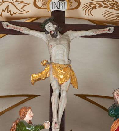 Zdjęcie nr 6: Grupa Ukrzyżowania złożona z ukrzyżowanego Chrystusa w typie christo vivo, stojących postaci Matki Boskiej i św. Jana oraz klęczącej Marii Magdaleny. Figura Chrystusa jest rzeźbą pełnoplastyczną, nieznacznie ściętą z tyłu, pozostałe są ścięte i wydrążone od tyłu. Zbawiciel przybity trzema gwoździami, z niemal prosto rozpostartymi rękami, z głową przechyloną na prawe ramię. Jego twarz jest pociągła, nos długi, oczy otwarte, twarz okolona kręconą, brązową brodą na głowie kręcone, sięgające szyi włosy i korona cierniowa pomalowana na zielono. Ręce z wyłamanymi barkami, z uwidocznionymi ścięgnami, klatka piersiowa wybrzuszona, z widocznymi żebrami i mostkiem, nogi smukłe z wyeksponowanymi kośćmi i mięśniami. Złocone perizonium przepasane podwójnym sznurem, z dwiema swobodnie rozwianymi połami po obu stronach bioder. Na naturalistycznie oddanym ciele zaznaczone strużki krwi. Na górnej belce titulus z literami: INRI". 
Matka Boska przedstawiona w pozycji stojącej, w kontrapoście, z rękami skrzyżowanymi na piersi. Głowę zwraca w kierunku Chrystusa, jej twarz jest owalna, włosy długie, brązowe, opadające na ramiona. Ubrana jest w czerwoną suknię z żółtą lamówką na rękawach, niebieski płaszcz o żółtym podbiciu i białą, długą chustę na głowie.    
Święty Jan ukazany w pozycji stojącej, w kontrapoście, z rękami złożonymi w geście modlitwy. Jego twarz jest owalna, oczy szeroko otwarte, włosy brązowe, kręcone, opadające na ramiona. Apostoł jest ubrany w zieloną tunikę i czerwony płaszcz z pomarańczowym podbiciem, przewieszony przez lewe ramię i ugiętą rękę. 
Po prawej stronie krzyża klęczy Maria Magdalena, ukazana prawym profilem, z prawą ręką, w której trzyma gąbkę wyciągniętą ku krzyżowi. Jej twarz jest pociągła z prostym nosem, włosy jasnobrązowe, długie, kręcone, opadające na plecy. Jest ubrana w zieloną tunikę i brązowy płaszcz o jaśniejszym podbiciu, opadający z lewego ramienia.
Na belce w kolorze czerwonym złocony napis: "Benedictus XVI SPE SALVI FACTI SUMUS A(NNO) D(OMINI) MMVII".
