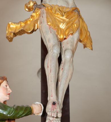 Zdjęcie nr 8: Grupa Ukrzyżowania złożona z ukrzyżowanego Chrystusa w typie christo vivo, stojących postaci Matki Boskiej i św. Jana oraz klęczącej Marii Magdaleny. Figura Chrystusa jest rzeźbą pełnoplastyczną, nieznacznie ściętą z tyłu, pozostałe są ścięte i wydrążone od tyłu. Zbawiciel przybity trzema gwoździami, z niemal prosto rozpostartymi rękami, z głową przechyloną na prawe ramię. Jego twarz jest pociągła, nos długi, oczy otwarte, twarz okolona kręconą, brązową brodą na głowie kręcone, sięgające szyi włosy i korona cierniowa pomalowana na zielono. Ręce z wyłamanymi barkami, z uwidocznionymi ścięgnami, klatka piersiowa wybrzuszona, z widocznymi żebrami i mostkiem, nogi smukłe z wyeksponowanymi kośćmi i mięśniami. Złocone perizonium przepasane podwójnym sznurem, z dwiema swobodnie rozwianymi połami po obu stronach bioder. Na naturalistycznie oddanym ciele zaznaczone strużki krwi. Na górnej belce titulus z literami: INRI". 
Matka Boska przedstawiona w pozycji stojącej, w kontrapoście, z rękami skrzyżowanymi na piersi. Głowę zwraca w kierunku Chrystusa, jej twarz jest owalna, włosy długie, brązowe, opadające na ramiona. Ubrana jest w czerwoną suknię z żółtą lamówką na rękawach, niebieski płaszcz o żółtym podbiciu i białą, długą chustę na głowie.    
Święty Jan ukazany w pozycji stojącej, w kontrapoście, z rękami złożonymi w geście modlitwy. Jego twarz jest owalna, oczy szeroko otwarte, włosy brązowe, kręcone, opadające na ramiona. Apostoł jest ubrany w zieloną tunikę i czerwony płaszcz z pomarańczowym podbiciem, przewieszony przez lewe ramię i ugiętą rękę. 
Po prawej stronie krzyża klęczy Maria Magdalena, ukazana prawym profilem, z prawą ręką, w której trzyma gąbkę wyciągniętą ku krzyżowi. Jej twarz jest pociągła z prostym nosem, włosy jasnobrązowe, długie, kręcone, opadające na plecy. Jest ubrana w zieloną tunikę i brązowy płaszcz o jaśniejszym podbiciu, opadający z lewego ramienia.
Na belce w kolorze czerwonym złocony napis: "Benedictus XVI SPE SALVI FACTI SUMUS A(NNO) D(OMINI) MMVII".
