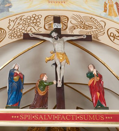 Zdjęcie nr 2: Grupa Ukrzyżowania złożona z ukrzyżowanego Chrystusa w typie christo vivo, stojących postaci Matki Boskiej i św. Jana oraz klęczącej Marii Magdaleny. Figura Chrystusa jest rzeźbą pełnoplastyczną, nieznacznie ściętą z tyłu, pozostałe są ścięte i wydrążone od tyłu. Zbawiciel przybity trzema gwoździami, z niemal prosto rozpostartymi rękami, z głową przechyloną na prawe ramię. Jego twarz jest pociągła, nos długi, oczy otwarte, twarz okolona kręconą, brązową brodą na głowie kręcone, sięgające szyi włosy i korona cierniowa pomalowana na zielono. Ręce z wyłamanymi barkami, z uwidocznionymi ścięgnami, klatka piersiowa wybrzuszona, z widocznymi żebrami i mostkiem, nogi smukłe z wyeksponowanymi kośćmi i mięśniami. Złocone perizonium przepasane podwójnym sznurem, z dwiema swobodnie rozwianymi połami po obu stronach bioder. Na naturalistycznie oddanym ciele zaznaczone strużki krwi. Na górnej belce titulus z literami: INRI". 
Matka Boska przedstawiona w pozycji stojącej, w kontrapoście, z rękami skrzyżowanymi na piersi. Głowę zwraca w kierunku Chrystusa, jej twarz jest owalna, włosy długie, brązowe, opadające na ramiona. Ubrana jest w czerwoną suknię z żółtą lamówką na rękawach, niebieski płaszcz o żółtym podbiciu i białą, długą chustę na głowie.    
Święty Jan ukazany w pozycji stojącej, w kontrapoście, z rękami złożonymi w geście modlitwy. Jego twarz jest owalna, oczy szeroko otwarte, włosy brązowe, kręcone, opadające na ramiona. Apostoł jest ubrany w zieloną tunikę i czerwony płaszcz z pomarańczowym podbiciem, przewieszony przez lewe ramię i ugiętą rękę. 
Po prawej stronie krzyża klęczy Maria Magdalena, ukazana prawym profilem, z prawą ręką, w której trzyma gąbkę wyciągniętą ku krzyżowi. Jej twarz jest pociągła z prostym nosem, włosy jasnobrązowe, długie, kręcone, opadające na plecy. Jest ubrana w zieloną tunikę i brązowy płaszcz o jaśniejszym podbiciu, opadający z lewego ramienia.
Na belce w kolorze czerwonym złocony napis: "Benedictus XVI SPE SALVI FACTI SUMUS A(NNO) D(OMINI) MMVII".
