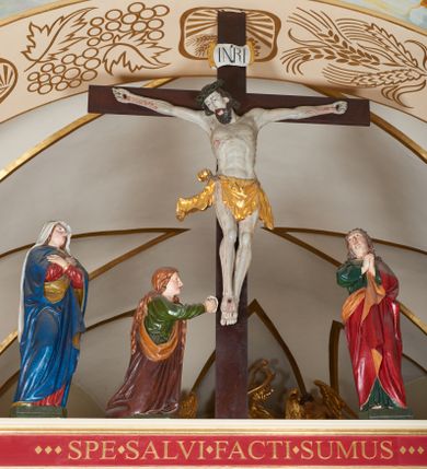 Zdjęcie nr 1: Grupa Ukrzyżowania złożona z ukrzyżowanego Chrystusa w typie christo vivo, stojących postaci Matki Boskiej i św. Jana oraz klęczącej Marii Magdaleny. Figura Chrystusa jest rzeźbą pełnoplastyczną, nieznacznie ściętą z tyłu, pozostałe są ścięte i wydrążone od tyłu. Zbawiciel przybity trzema gwoździami, z niemal prosto rozpostartymi rękami, z głową przechyloną na prawe ramię. Jego twarz jest pociągła, nos długi, oczy otwarte, twarz okolona kręconą, brązową brodą na głowie kręcone, sięgające szyi włosy i korona cierniowa pomalowana na zielono. Ręce z wyłamanymi barkami, z uwidocznionymi ścięgnami, klatka piersiowa wybrzuszona, z widocznymi żebrami i mostkiem, nogi smukłe z wyeksponowanymi kośćmi i mięśniami. Złocone perizonium przepasane podwójnym sznurem, z dwiema swobodnie rozwianymi połami po obu stronach bioder. Na naturalistycznie oddanym ciele zaznaczone strużki krwi. Na górnej belce titulus z literami: INRI". 
Matka Boska przedstawiona w pozycji stojącej, w kontrapoście, z rękami skrzyżowanymi na piersi. Głowę zwraca w kierunku Chrystusa, jej twarz jest owalna, włosy długie, brązowe, opadające na ramiona. Ubrana jest w czerwoną suknię z żółtą lamówką na rękawach, niebieski płaszcz o żółtym podbiciu i białą, długą chustę na głowie.    
Święty Jan ukazany w pozycji stojącej, w kontrapoście, z rękami złożonymi w geście modlitwy. Jego twarz jest owalna, oczy szeroko otwarte, włosy brązowe, kręcone, opadające na ramiona. Apostoł jest ubrany w zieloną tunikę i czerwony płaszcz z pomarańczowym podbiciem, przewieszony przez lewe ramię i ugiętą rękę. 
Po prawej stronie krzyża klęczy Maria Magdalena, ukazana prawym profilem, z prawą ręką, w której trzyma gąbkę wyciągniętą ku krzyżowi. Jej twarz jest pociągła z prostym nosem, włosy jasnobrązowe, długie, kręcone, opadające na plecy. Jest ubrana w zieloną tunikę i brązowy płaszcz o jaśniejszym podbiciu, opadający z lewego ramienia.
Na belce w kolorze czerwonym złocony napis: "Benedictus XVI SPE SALVI FACTI SUMUS A(NNO) D(OMINI) MMVII".
