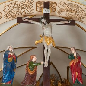 Zdjęcie nr 1: Grupa Ukrzyżowania złożona z ukrzyżowanego Chrystusa w typie christo vivo, stojących postaci Matki Boskiej i św. Jana oraz klęczącej Marii Magdaleny. Figura Chrystusa jest rzeźbą pełnoplastyczną, nieznacznie ściętą z tyłu, pozostałe są ścięte i wydrążone od tyłu. Zbawiciel przybity trzema gwoździami, z niemal prosto rozpostartymi rękami, z głową przechyloną na prawe ramię. Jego twarz jest pociągła, nos długi, oczy otwarte, twarz okolona kręconą, brązową brodą na głowie kręcone, sięgające szyi włosy i korona cierniowa pomalowana na zielono. Ręce z wyłamanymi barkami, z uwidocznionymi ścięgnami, klatka piersiowa wybrzuszona, z widocznymi żebrami i mostkiem, nogi smukłe z wyeksponowanymi kośćmi i mięśniami. Złocone perizonium przepasane podwójnym sznurem, z dwiema swobodnie rozwianymi połami po obu stronach bioder. Na naturalistycznie oddanym ciele zaznaczone strużki krwi. Na górnej belce titulus z literami: INRI&quot;. 
Matka Boska przedstawiona w pozycji stojącej, w kontrapoście, z rękami skrzyżowanymi na piersi. Głowę zwraca w kierunku Chrystusa, jej twarz jest owalna, włosy długie, brązowe, opadające na ramiona. Ubrana jest w czerwoną suknię z żółtą lamówką na rękawach, niebieski płaszcz o żółtym podbiciu i białą, długą chustę na głowie.    
Święty Jan ukazany w pozycji stojącej, w kontrapoście, z rękami złożonymi w geście modlitwy. Jego twarz jest owalna, oczy szeroko otwarte, włosy brązowe, kręcone, opadające na ramiona. Apostoł jest ubrany w zieloną tunikę i czerwony płaszcz z pomarańczowym podbiciem, przewieszony przez lewe ramię i ugiętą rękę. 
Po prawej stronie krzyża klęczy Maria Magdalena, ukazana prawym profilem, z prawą ręką, w której trzyma gąbkę wyciągniętą ku krzyżowi. Jej twarz jest pociągła z prostym nosem, włosy jasnobrązowe, długie, kręcone, opadające na plecy. Jest ubrana w zieloną tunikę i brązowy płaszcz o jaśniejszym podbiciu, opadający z lewego ramienia.
Na belce w kolorze czerwonym złocony napis: &quot;Benedictus XVI SPE SALVI FACTI SUMUS A(NNO) D(OMINI) MMVII&quot;.
