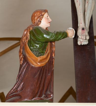 Zdjęcie nr 12: Grupa Ukrzyżowania złożona z ukrzyżowanego Chrystusa w typie christo vivo, stojących postaci Matki Boskiej i św. Jana oraz klęczącej Marii Magdaleny. Figura Chrystusa jest rzeźbą pełnoplastyczną, nieznacznie ściętą z tyłu, pozostałe są ścięte i wydrążone od tyłu. Zbawiciel przybity trzema gwoździami, z niemal prosto rozpostartymi rękami, z głową przechyloną na prawe ramię. Jego twarz jest pociągła, nos długi, oczy otwarte, twarz okolona kręconą, brązową brodą na głowie kręcone, sięgające szyi włosy i korona cierniowa pomalowana na zielono. Ręce z wyłamanymi barkami, z uwidocznionymi ścięgnami, klatka piersiowa wybrzuszona, z widocznymi żebrami i mostkiem, nogi smukłe z wyeksponowanymi kośćmi i mięśniami. Złocone perizonium przepasane podwójnym sznurem, z dwiema swobodnie rozwianymi połami po obu stronach bioder. Na naturalistycznie oddanym ciele zaznaczone strużki krwi. Na górnej belce titulus z literami: INRI". 
Matka Boska przedstawiona w pozycji stojącej, w kontrapoście, z rękami skrzyżowanymi na piersi. Głowę zwraca w kierunku Chrystusa, jej twarz jest owalna, włosy długie, brązowe, opadające na ramiona. Ubrana jest w czerwoną suknię z żółtą lamówką na rękawach, niebieski płaszcz o żółtym podbiciu i białą, długą chustę na głowie.    
Święty Jan ukazany w pozycji stojącej, w kontrapoście, z rękami złożonymi w geście modlitwy. Jego twarz jest owalna, oczy szeroko otwarte, włosy brązowe, kręcone, opadające na ramiona. Apostoł jest ubrany w zieloną tunikę i czerwony płaszcz z pomarańczowym podbiciem, przewieszony przez lewe ramię i ugiętą rękę. 
Po prawej stronie krzyża klęczy Maria Magdalena, ukazana prawym profilem, z prawą ręką, w której trzyma gąbkę wyciągniętą ku krzyżowi. Jej twarz jest pociągła z prostym nosem, włosy jasnobrązowe, długie, kręcone, opadające na plecy. Jest ubrana w zieloną tunikę i brązowy płaszcz o jaśniejszym podbiciu, opadający z lewego ramienia.
Na belce w kolorze czerwonym złocony napis: "Benedictus XVI SPE SALVI FACTI SUMUS A(NNO) D(OMINI) MMVII".
