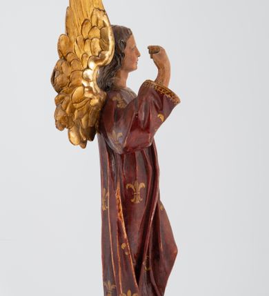 Zdjęcie nr 3: Pełnoplastyczna rzeźba stojącego w kontrapoście na chmurach anioła, z głową zwróconą w lewo, z prawą ręką, ugiętą w łokciu, wzniesioną wzwyż, lewą opuszczoną wzdłuż ciała. Twarz o rysach młodzieńca z prostym nosem, różowymi ustami, niebieskimi oczami, okolona włosami opadającymi w lokach na plecy. Anioł ubrany w długą czerwoną tunikę z szerokimi rękawami, malowaną w złote lilie herbowe. Strzeliste skrzydła rozpostarte, malowane na złoto. 