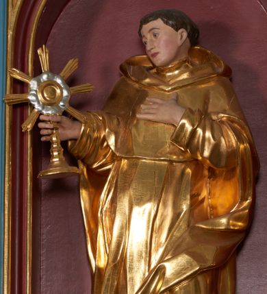 Zdjęcie nr 1: Rzeźba przyścienna, całopostaciowa, zapewne drążona od tyłu. Święty jest ujęty w lekkim kontrapoście, z ugiętą lewą nogą. Jego lewa ręka jest zgięta w łokciu i złożona na piersi, w prawej trzyma monstrancję. Głowa jest zwrócona w prawo i lekko pochylona, twarz o ostrych rysach, prostym nosie, małych oczach. Na głowie tonsura i brązowe, krótkie włosy. Święty jest ubrany w habit dominikański i płaszcz z kapturem zarzucony na lewe kolano, spod habitu wystają brązowe buty. Figura polichromowana naturalistycznie w partiach ciała ze złoconym strojem oraz złoconą i srebrzoną monstrancją.
