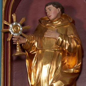 Zdjęcie nr 1: Rzeźba przyścienna, całopostaciowa, zapewne drążona od tyłu. Święty jest ujęty w lekkim kontrapoście, z ugiętą lewą nogą. Jego lewa ręka jest zgięta w łokciu i złożona na piersi, w prawej trzyma monstrancję. Głowa jest zwrócona w prawo i lekko pochylona, twarz o ostrych rysach, prostym nosie, małych oczach. Na głowie tonsura i brązowe, krótkie włosy. Święty jest ubrany w habit dominikański i płaszcz z kapturem zarzucony na lewe kolano, spod habitu wystają brązowe buty. Figura polichromowana naturalistycznie w partiach ciała ze złoconym strojem oraz złoconą i srebrzoną monstrancją.
