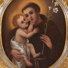 Zdjęcie nr 1: Obraz ujęty ramą w kształcie stojącego prostokąta z wycięciem w kształcie stojącego owalu, profilowaną, zdobioną motywami roślinno-kwiatowymi w narożach oraz wicią suchego akantu. Święty Antoni ujęty półpostaciowo, w trzech czwartych zwrócony w prawo. Jego twarz jest owalna o delikatnych, młodzieńczych rysach. Włosy ma krótkie, ciemnobrązowe, z wyciętą tonsurą. Święty jest ubrany w brązowy habit franciszkański. W lewej dłoni trzyma białą lilię, prawą podtrzymuje Dzieciątko Jezus. Trzymany na ręku Jezus ukazany w pozycji siedzącej, w trzech czwartych, zwrócony w lewo. Jego głowa jest odchylona do tyłu, wzrok skierowany w stronę twarzy świętego. Twarz ma owalną o delikatnych rysach, włosy jasnobrązowe, krótkie i falowane. Jego biodra i częściowo nóżki przesłania biała pielucha. Lewą rączkę wspiera na podbródku świętego, prawą unosi na wysokości klatki piersiowej. Tło nieokreślone, brązowe, rozświetlone nimbami wokół głów postaci. Kolorystyka w ciepłej tonacji, nasycona, o delikatnym modelunku.  
