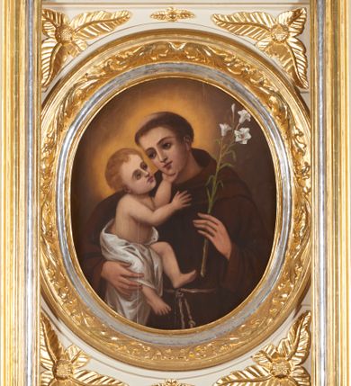 Zdjęcie nr 2: Obraz ujęty ramą w kształcie stojącego prostokąta z wycięciem w kształcie stojącego owalu, profilowaną, zdobioną motywami roślinno-kwiatowymi w narożach oraz wicią suchego akantu. Święty Antoni ujęty półpostaciowo, w trzech czwartych zwrócony w prawo. Jego twarz jest owalna o delikatnych, młodzieńczych rysach. Włosy ma krótkie, ciemnobrązowe, z wyciętą tonsurą. Święty jest ubrany w brązowy habit franciszkański. W lewej dłoni trzyma białą lilię, prawą podtrzymuje Dzieciątko Jezus. Trzymany na ręku Jezus ukazany w pozycji siedzącej, w trzech czwartych, zwrócony w lewo. Jego głowa jest odchylona do tyłu, wzrok skierowany w stronę twarzy świętego. Twarz ma owalną o delikatnych rysach, włosy jasnobrązowe, krótkie i falowane. Jego biodra i częściowo nóżki przesłania biała pielucha. Lewą rączkę wspiera na podbródku świętego, prawą unosi na wysokości klatki piersiowej. Tło nieokreślone, brązowe, rozświetlone nimbami wokół głów postaci. Kolorystyka w ciepłej tonacji, nasycona, o delikatnym modelunku.  
