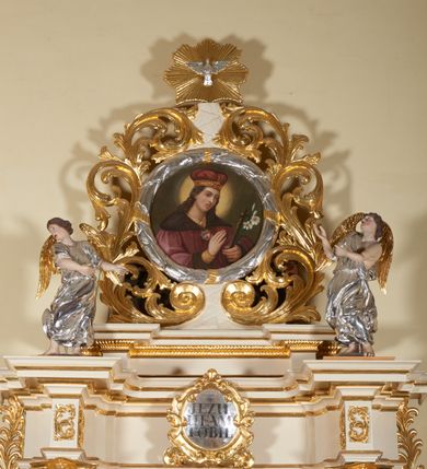 Zdjęcie nr 7: Figury pełnoplastyczne, wydrążone od tyłu, ukazują stojące w zwieńczeniu ołtarza anioły. Figura po lewej stronie jest zwrócona w lewo, o wygiętej łukowo sylwetce, z głową skręconą w prawo. Jej twarz jest owalna, o wyrazistych rysach i mimice oraz jasnej, szarawej karnacji z zaróżowionymi policzkami. Włosy ma krótkie, zaczesane do tyłu, falowane, ciemnobrązowe. Ręce uniesione na wysokości bioder i wysunięte równolegle do przodu. Anioł jest ubrany w długą, podwiązaną w pasie, mocno sfałdowaną, srebrzoną tunikę z rękawami sięgającymi łokci, z podwiniętym dołem. Na plecach smukłe, złocone skrzydła. Figura po prawej stronie jest zwrócona w prawo, ujęta w kontrapoście, o przechylonej do tyłu sylwetce. Jej głowa jest lekko odchylona do tyłu, twarz owalna o wyrazistych rysach, szarawej karnacji z zaróżowionymi policzkami. Włosy krótkie, zaczesane do tyłu, falowane, ciemnobrązowe. Ręce wysunięte przed korpus, lekko uniesione na wysokości klatki piersiowej. Anioł jest ubrany w długą, podwiązaną w pasie, mocno sfałdowaną, srebrzoną tunikę z rękawami sięgającymi łokci, z odsłoniętym lewym ramieniem i ręką. Na plecach smukłe, złocone skrzydła. 


