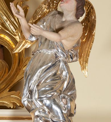 Zdjęcie nr 5: Figury pełnoplastyczne, wydrążone od tyłu, ukazują stojące w zwieńczeniu ołtarza anioły. Figura po lewej stronie jest zwrócona w lewo, o wygiętej łukowo sylwetce, z głową skręconą w prawo. Jej twarz jest owalna, o wyrazistych rysach i mimice oraz jasnej, szarawej karnacji z zaróżowionymi policzkami. Włosy ma krótkie, zaczesane do tyłu, falowane, ciemnobrązowe. Ręce uniesione na wysokości bioder i wysunięte równolegle do przodu. Anioł jest ubrany w długą, podwiązaną w pasie, mocno sfałdowaną, srebrzoną tunikę z rękawami sięgającymi łokci, z podwiniętym dołem. Na plecach smukłe, złocone skrzydła. Figura po prawej stronie jest zwrócona w prawo, ujęta w kontrapoście, o przechylonej do tyłu sylwetce. Jej głowa jest lekko odchylona do tyłu, twarz owalna o wyrazistych rysach, szarawej karnacji z zaróżowionymi policzkami. Włosy krótkie, zaczesane do tyłu, falowane, ciemnobrązowe. Ręce wysunięte przed korpus, lekko uniesione na wysokości klatki piersiowej. Anioł jest ubrany w długą, podwiązaną w pasie, mocno sfałdowaną, srebrzoną tunikę z rękawami sięgającymi łokci, z odsłoniętym lewym ramieniem i ręką. Na plecach smukłe, złocone skrzydła. 

