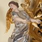 Zdjęcie nr 1: Figury pełnoplastyczne, wydrążone od tyłu, ukazują stojące w zwieńczeniu ołtarza anioły. Figura po lewej stronie jest zwrócona w lewo, o wygiętej łukowo sylwetce, z głową skręconą w prawo. Jej twarz jest owalna, o wyrazistych rysach i mimice oraz jasnej, szarawej karnacji z zaróżowionymi policzkami. Włosy ma krótkie, zaczesane do tyłu, falowane, ciemnobrązowe. Ręce uniesione na wysokości bioder i wysunięte równolegle do przodu. Anioł jest ubrany w długą, podwiązaną w pasie, mocno sfałdowaną, srebrzoną tunikę z rękawami sięgającymi łokci, z podwiniętym dołem. Na plecach smukłe, złocone skrzydła. Figura po prawej stronie jest zwrócona w prawo, ujęta w kontrapoście, o przechylonej do tyłu sylwetce. Jej głowa jest lekko odchylona do tyłu, twarz owalna o wyrazistych rysach, szarawej karnacji z zaróżowionymi policzkami. Włosy krótkie, zaczesane do tyłu, falowane, ciemnobrązowe. Ręce wysunięte przed korpus, lekko uniesione na wysokości klatki piersiowej. Anioł jest ubrany w długą, podwiązaną w pasie, mocno sfałdowaną, srebrzoną tunikę z rękawami sięgającymi łokci, z odsłoniętym lewym ramieniem i ręką. Na plecach smukłe, złocone skrzydła. 

