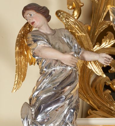 Zdjęcie nr 1: Figury pełnoplastyczne, wydrążone od tyłu, ukazują stojące w zwieńczeniu ołtarza anioły. Figura po lewej stronie jest zwrócona w lewo, o wygiętej łukowo sylwetce, z głową skręconą w prawo. Jej twarz jest owalna, o wyrazistych rysach i mimice oraz jasnej, szarawej karnacji z zaróżowionymi policzkami. Włosy ma krótkie, zaczesane do tyłu, falowane, ciemnobrązowe. Ręce uniesione na wysokości bioder i wysunięte równolegle do przodu. Anioł jest ubrany w długą, podwiązaną w pasie, mocno sfałdowaną, srebrzoną tunikę z rękawami sięgającymi łokci, z podwiniętym dołem. Na plecach smukłe, złocone skrzydła. Figura po prawej stronie jest zwrócona w prawo, ujęta w kontrapoście, o przechylonej do tyłu sylwetce. Jej głowa jest lekko odchylona do tyłu, twarz owalna o wyrazistych rysach, szarawej karnacji z zaróżowionymi policzkami. Włosy krótkie, zaczesane do tyłu, falowane, ciemnobrązowe. Ręce wysunięte przed korpus, lekko uniesione na wysokości klatki piersiowej. Anioł jest ubrany w długą, podwiązaną w pasie, mocno sfałdowaną, srebrzoną tunikę z rękawami sięgającymi łokci, z odsłoniętym lewym ramieniem i ręką. Na plecach smukłe, złocone skrzydła. 

