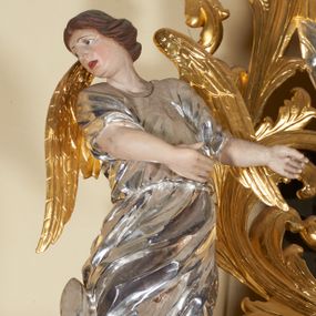 Zdjęcie nr 1: Figury pełnoplastyczne, wydrążone od tyłu, ukazują stojące w zwieńczeniu ołtarza anioły. Figura po lewej stronie jest zwrócona w lewo, o wygiętej łukowo sylwetce, z głową skręconą w prawo. Jej twarz jest owalna, o wyrazistych rysach i mimice oraz jasnej, szarawej karnacji z zaróżowionymi policzkami. Włosy ma krótkie, zaczesane do tyłu, falowane, ciemnobrązowe. Ręce uniesione na wysokości bioder i wysunięte równolegle do przodu. Anioł jest ubrany w długą, podwiązaną w pasie, mocno sfałdowaną, srebrzoną tunikę z rękawami sięgającymi łokci, z podwiniętym dołem. Na plecach smukłe, złocone skrzydła. Figura po prawej stronie jest zwrócona w prawo, ujęta w kontrapoście, o przechylonej do tyłu sylwetce. Jej głowa jest lekko odchylona do tyłu, twarz owalna o wyrazistych rysach, szarawej karnacji z zaróżowionymi policzkami. Włosy krótkie, zaczesane do tyłu, falowane, ciemnobrązowe. Ręce wysunięte przed korpus, lekko uniesione na wysokości klatki piersiowej. Anioł jest ubrany w długą, podwiązaną w pasie, mocno sfałdowaną, srebrzoną tunikę z rękawami sięgającymi łokci, z odsłoniętym lewym ramieniem i ręką. Na plecach smukłe, złocone skrzydła. 

