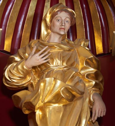 Zdjęcie nr 1: Święta Anna została ukazana frontalnie, całopostaciowo, w lekkim kontrapoście, z prawą ręką zgiętą w łokciu i uniesioną na wysokości piersi, a lewą opuszczoną wzdłuż ciała. Ubrana w długą suknię przewiązaną, płaszcz zarzucony na plecy i welon na głowie. Ma twarz o ostrych rysach, z wydatnymi kościami policzkowymi, szerokim, dużym nosem, otwartymi oczami i zamkniętymi ustami. 