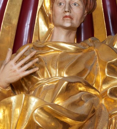 Zdjęcie nr 5: Święta Anna została ukazana frontalnie, całopostaciowo, w lekkim kontrapoście, z prawą ręką zgiętą w łokciu i uniesioną na wysokości piersi, a lewą opuszczoną wzdłuż ciała. Ubrana w długą suknię przewiązaną, płaszcz zarzucony na plecy i welon na głowie. Ma twarz o ostrych rysach, z wydatnymi kościami policzkowymi, szerokim, dużym nosem, otwartymi oczami i zamkniętymi ustami. 