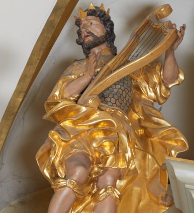 Zdjęcie nr 1: Figura całopostaciowa, zwrócona frontalnie, w kontrapoście, delikatnie przechylona w prawo, z harfą w rękach wspartą na biodrze. Król Dawid jest ubrany w zbroję łuskową, z krótkimi piórami i płaszcz narzucony na ramiona, opadający na plecy z zawiniętym fragmentem na prawej nodze. Jego twarz jest owalna, nos niewielki, delikatnie zadarty, oczy małe, wpatrzone na wprost, brwi uniesione, usta pełne, rozchylone; okolona krótką brodą. Włosy krótkie, silnie skręcone w poszczególne pasma loków okalających twarz. Tkanina jego ubioru ekspresyjnie drapowana w płynne, miękkie fałdy, miejscami skręcona, z rozwianym fragmentem przy lewym boku. Na głowie Dawida korona otwarta. Pod kolanami zawiązane wstęgi, na stopach widoczne sandały. 