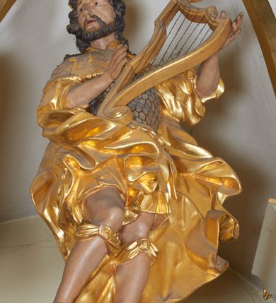 Zdjęcie nr 2: Figura całopostaciowa, zwrócona frontalnie, w kontrapoście, delikatnie przechylona w prawo, z harfą w rękach wspartą na biodrze. Król Dawid jest ubrany w zbroję łuskową, z krótkimi piórami i płaszcz narzucony na ramiona, opadający na plecy z zawiniętym fragmentem na prawej nodze. Jego twarz jest owalna, nos niewielki, delikatnie zadarty, oczy małe, wpatrzone na wprost, brwi uniesione, usta pełne, rozchylone; okolona krótką brodą. Włosy krótkie, silnie skręcone w poszczególne pasma loków okalających twarz. Tkanina jego ubioru ekspresyjnie drapowana w płynne, miękkie fałdy, miejscami skręcona, z rozwianym fragmentem przy lewym boku. Na głowie Dawida korona otwarta. Pod kolanami zawiązane wstęgi, na stopach widoczne sandały. 