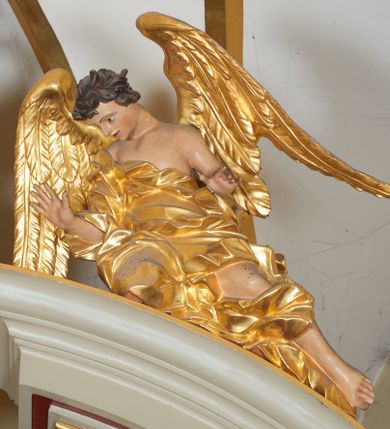 Zdjęcie nr 2: Para figur aniołów na przerwanym przyczółku w układzie przeciwstawnym. Ukazane w pozycji siedzącej, z podkurczoną prawą i wyciągniętymi wzdłuż przyczółka nogami, z pochyloną głową i rozłożonymi rękami. Ich twarze są szczupłe, o wyrazistych rysach, otwartych oczach i ustach. Włosy krótkie, rzeźbione w pojedyncze silnie skręcone, ekspresyjne pukle. Anioły ubrane są w szaty przewiązane przez jedno ramię, odsłaniające tors i fragmenty nóg, modelowane obficie w liczne fałdy i marszczenia. Skrzydła duże, delikatnie rozłożone. Polichromia naturalistyczna w odsłoniętych partiach ciała, szaty i atrybuty złocone.
