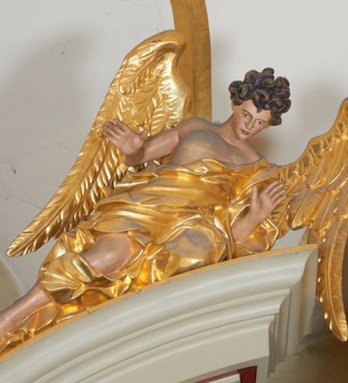 Zdjęcie nr 1: Para figur aniołów na przerwanym przyczółku w układzie przeciwstawnym. Ukazane w pozycji siedzącej, z podkurczoną prawą i wyciągniętymi wzdłuż przyczółka nogami, z pochyloną głową i rozłożonymi rękami. Ich twarze są szczupłe, o wyrazistych rysach, otwartych oczach i ustach. Włosy krótkie, rzeźbione w pojedyncze silnie skręcone, ekspresyjne pukle. Anioły ubrane są w szaty przewiązane przez jedno ramię, odsłaniające tors i fragmenty nóg, modelowane obficie w liczne fałdy i marszczenia. Skrzydła duże, delikatnie rozłożone. Polichromia naturalistyczna w odsłoniętych partiach ciała, szaty i atrybuty złocone.
