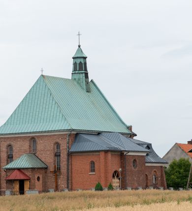 Zdjęcie nr 42: Kościół murowany z cegły na niskiej, kamiennej podmurówce, orientowany, jednonawowy ze zredukowanym transeptem oraz nieco niższym, zamkniętym półkoliście prezbiterium. Do północno-wschodniego narożnika nawy (przy prezbiterium) przylega założona na rzucie prostokąta, oszkarpowana zakrystia dostępna półkolistym portalem od strony północnej. Do południowo-wschodniego narożnika nawy dostawiona kaplica Matki Boskiej na rzucie prostokąta zamknięta od wschodu niewielką absydą. Ramiona transeptu zredukowane do roli prostokątnych kaplic. Do południowo-zachodniego i północno-zachodniego narożnika nawy dobudowane nowsze kruchty boczne założone na rzucie prostokąta dostępne portalami o półkolistym wykroju. Kruchta zachodnia na rzucie prostokąta dostępna nowym, prostokątnym wejściem. W ścianach północnej i południowej widoczne, zamurowane, starsze otwory wejściowe (od południa w miejsce portalu okrągłe okno). Dach nad nawą dwuspadowy z trzecią połacią zakrywającą ścianę szczytową; w części wschodniej wieloboczna wieżyczka na sygnaturkę z glorietą zwieńczoną krzyżem. Kruchty oraz kaplice boczne kryte dachami trójpołaciowymi, prezbiterium jednopołaciowym; nad zakrystią dach mansardowy z prostokątnym oknem w lukarnie. Pokrycia dachowe wykonane są z blachy (pierwotnie była to czerwona dachówka). W nawie i prezbiterium strop o konstrukcji żelbetowej wsparty na dziewięciu masywnych kolumnach ustawionych na wysokich cokołach; kaplice boczne sklepione krzyżowo, w zakrystii założony betonowy strop. Chór muzyczny murowany, wsparty na pierwszej od zachodu parze kolumn dźwigających strop. Fasada jednokondygnacyjna, trójosiowa z umieszczoną w osi centralnej prostopadłościenną kruchtą mieszczącą prostokątny otwór wejściowy (nowy). Okna w kształcie stojącego prostokąta o smukłych proporcjach, zamknięte półkoliście. Okno środkowe, umieszczone nieco wyżej, niższe od pozostałych. W ścianach zachodnich krucht bocznych po jednym niewielkim oknie o wykroju półkolistym. Trójkątna ściana szczytowa zasłonięta blaszanym dachem o przełamanej w dolnej części połaci, zwieńczona niewielkim, metalowym krzyżem. Elewacje boczne jednokondygnacyjne, o nieregularnym przebiegu osi. W elewacji południowej w pierwszej osi ceglany portal z nadświetlem o wykroju półkolistym, w drugiej okrągłe okno doświetlające kaplicę boczną, w kolejnych: półkolisty ceglany portal do zakrystii oraz okno o półkolistym wykroju z motywem biforium. W elewacji północnej w osi pierwszej i drugiej niewielkie półkoliście wykrojone okna, w osi trzeciej okrągłe okno doświetlające kaplicę, w osi czwartej natomiast ceglany portal  z nadświetlem o wykroju półkolistym. W prezbiterium cztery okna o wykroju półkolistym. W trzeciej osi, pomiędzy oknami, ujęta wielobocznymi filarami dźwigającymi, wydatny, profilowany gzyms tynkowana na biało nisza z figurą Matki Boskiej Fatimskiej umieszczonej pod baldachimem. Górną część murów nawy, prezbiterium, zakrystii, kaplic oraz obu krucht bocznych obiega profilowany gzyms okapowy. 