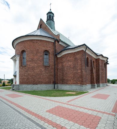 Zdjęcie nr 8: Kościół murowany z cegły na niskiej, kamiennej podmurówce, orientowany, jednonawowy ze zredukowanym transeptem oraz nieco niższym, zamkniętym półkoliście prezbiterium. Do północno-wschodniego narożnika nawy (przy prezbiterium) przylega założona na rzucie prostokąta, oszkarpowana zakrystia dostępna półkolistym portalem od strony północnej. Do południowo-wschodniego narożnika nawy dostawiona kaplica Matki Boskiej na rzucie prostokąta zamknięta od wschodu niewielką absydą. Ramiona transeptu zredukowane do roli prostokątnych kaplic. Do południowo-zachodniego i północno-zachodniego narożnika nawy dobudowane nowsze kruchty boczne założone na rzucie prostokąta dostępne portalami o półkolistym wykroju. Kruchta zachodnia na rzucie prostokąta dostępna nowym, prostokątnym wejściem. W ścianach północnej i południowej widoczne, zamurowane, starsze otwory wejściowe (od południa w miejsce portalu okrągłe okno). Dach nad nawą dwuspadowy z trzecią połacią zakrywającą ścianę szczytową; w części wschodniej wieloboczna wieżyczka na sygnaturkę z glorietą zwieńczoną krzyżem. Kruchty oraz kaplice boczne kryte dachami trójpołaciowymi, prezbiterium jednopołaciowym; nad zakrystią dach mansardowy z prostokątnym oknem w lukarnie. Pokrycia dachowe wykonane są z blachy (pierwotnie była to czerwona dachówka). W nawie i prezbiterium strop o konstrukcji żelbetowej wsparty na dziewięciu masywnych kolumnach ustawionych na wysokich cokołach; kaplice boczne sklepione krzyżowo, w zakrystii założony betonowy strop. Chór muzyczny murowany, wsparty na pierwszej od zachodu parze kolumn dźwigających strop. Fasada jednokondygnacyjna, trójosiowa z umieszczoną w osi centralnej prostopadłościenną kruchtą mieszczącą prostokątny otwór wejściowy (nowy). Okna w kształcie stojącego prostokąta o smukłych proporcjach, zamknięte półkoliście. Okno środkowe, umieszczone nieco wyżej, niższe od pozostałych. W ścianach zachodnich krucht bocznych po jednym niewielkim oknie o wykroju półkolistym. Trójkątna ściana szczytowa zasłonięta blaszanym dachem o przełamanej w dolnej części połaci, zwieńczona niewielkim, metalowym krzyżem. Elewacje boczne jednokondygnacyjne, o nieregularnym przebiegu osi. W elewacji południowej w pierwszej osi ceglany portal z nadświetlem o wykroju półkolistym, w drugiej okrągłe okno doświetlające kaplicę boczną, w kolejnych: półkolisty ceglany portal do zakrystii oraz okno o półkolistym wykroju z motywem biforium. W elewacji północnej w osi pierwszej i drugiej niewielkie półkoliście wykrojone okna, w osi trzeciej okrągłe okno doświetlające kaplicę, w osi czwartej natomiast ceglany portal  z nadświetlem o wykroju półkolistym. W prezbiterium cztery okna o wykroju półkolistym. W trzeciej osi, pomiędzy oknami, ujęta wielobocznymi filarami dźwigającymi, wydatny, profilowany gzyms tynkowana na biało nisza z figurą Matki Boskiej Fatimskiej umieszczonej pod baldachimem. Górną część murów nawy, prezbiterium, zakrystii, kaplic oraz obu krucht bocznych obiega profilowany gzyms okapowy. 