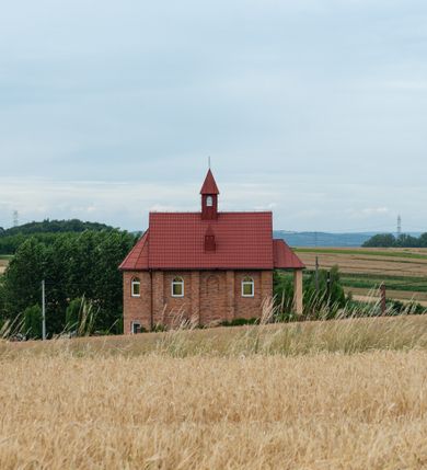 Zdjęcie nr 11: Kościół murowany z cegły na niskiej, kamiennej podmurówce, orientowany, jednonawowy ze zredukowanym transeptem oraz nieco niższym, zamkniętym półkoliście prezbiterium. Do północno-wschodniego narożnika nawy (przy prezbiterium) przylega założona na rzucie prostokąta, oszkarpowana zakrystia dostępna półkolistym portalem od strony północnej. Do południowo-wschodniego narożnika nawy dostawiona kaplica Matki Boskiej na rzucie prostokąta zamknięta od wschodu niewielką absydą. Ramiona transeptu zredukowane do roli prostokątnych kaplic. Do południowo-zachodniego i północno-zachodniego narożnika nawy dobudowane nowsze kruchty boczne założone na rzucie prostokąta dostępne portalami o półkolistym wykroju. Kruchta zachodnia na rzucie prostokąta dostępna nowym, prostokątnym wejściem. W ścianach północnej i południowej widoczne, zamurowane, starsze otwory wejściowe (od południa w miejsce portalu okrągłe okno). Dach nad nawą dwuspadowy z trzecią połacią zakrywającą ścianę szczytową; w części wschodniej wieloboczna wieżyczka na sygnaturkę z glorietą zwieńczoną krzyżem. Kruchty oraz kaplice boczne kryte dachami trójpołaciowymi, prezbiterium jednopołaciowym; nad zakrystią dach mansardowy z prostokątnym oknem w lukarnie. Pokrycia dachowe wykonane są z blachy (pierwotnie była to czerwona dachówka). W nawie i prezbiterium strop o konstrukcji żelbetowej wsparty na dziewięciu masywnych kolumnach ustawionych na wysokich cokołach; kaplice boczne sklepione krzyżowo, w zakrystii założony betonowy strop. Chór muzyczny murowany, wsparty na pierwszej od zachodu parze kolumn dźwigających strop. Fasada jednokondygnacyjna, trójosiowa z umieszczoną w osi centralnej prostopadłościenną kruchtą mieszczącą prostokątny otwór wejściowy (nowy). Okna w kształcie stojącego prostokąta o smukłych proporcjach, zamknięte półkoliście. Okno środkowe, umieszczone nieco wyżej, niższe od pozostałych. W ścianach zachodnich krucht bocznych po jednym niewielkim oknie o wykroju półkolistym. Trójkątna ściana szczytowa zasłonięta blaszanym dachem o przełamanej w dolnej części połaci, zwieńczona niewielkim, metalowym krzyżem. Elewacje boczne jednokondygnacyjne, o nieregularnym przebiegu osi. W elewacji południowej w pierwszej osi ceglany portal z nadświetlem o wykroju półkolistym, w drugiej okrągłe okno doświetlające kaplicę boczną, w kolejnych: półkolisty ceglany portal do zakrystii oraz okno o półkolistym wykroju z motywem biforium. W elewacji północnej w osi pierwszej i drugiej niewielkie półkoliście wykrojone okna, w osi trzeciej okrągłe okno doświetlające kaplicę, w osi czwartej natomiast ceglany portal  z nadświetlem o wykroju półkolistym. W prezbiterium cztery okna o wykroju półkolistym. W trzeciej osi, pomiędzy oknami, ujęta wielobocznymi filarami dźwigającymi, wydatny, profilowany gzyms tynkowana na biało nisza z figurą Matki Boskiej Fatimskiej umieszczonej pod baldachimem. Górną część murów nawy, prezbiterium, zakrystii, kaplic oraz obu krucht bocznych obiega profilowany gzyms okapowy. 