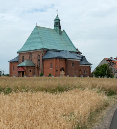 Zdjęcie nr 13: Kościół murowany z cegły na niskiej, kamiennej podmurówce, orientowany, jednonawowy ze zredukowanym transeptem oraz nieco niższym, zamkniętym półkoliście prezbiterium. Do północno-wschodniego narożnika nawy (przy prezbiterium) przylega założona na rzucie prostokąta, oszkarpowana zakrystia dostępna półkolistym portalem od strony północnej. Do południowo-wschodniego narożnika nawy dostawiona kaplica Matki Boskiej na rzucie prostokąta zamknięta od wschodu niewielką absydą. Ramiona transeptu zredukowane do roli prostokątnych kaplic. Do południowo-zachodniego i północno-zachodniego narożnika nawy dobudowane nowsze kruchty boczne założone na rzucie prostokąta dostępne portalami o półkolistym wykroju. Kruchta zachodnia na rzucie prostokąta dostępna nowym, prostokątnym wejściem. W ścianach północnej i południowej widoczne, zamurowane, starsze otwory wejściowe (od południa w miejsce portalu okrągłe okno). Dach nad nawą dwuspadowy z trzecią połacią zakrywającą ścianę szczytową; w części wschodniej wieloboczna wieżyczka na sygnaturkę z glorietą zwieńczoną krzyżem. Kruchty oraz kaplice boczne kryte dachami trójpołaciowymi, prezbiterium jednopołaciowym; nad zakrystią dach mansardowy z prostokątnym oknem w lukarnie. Pokrycia dachowe wykonane są z blachy (pierwotnie była to czerwona dachówka). W nawie i prezbiterium strop o konstrukcji żelbetowej wsparty na dziewięciu masywnych kolumnach ustawionych na wysokich cokołach; kaplice boczne sklepione krzyżowo, w zakrystii założony betonowy strop. Chór muzyczny murowany, wsparty na pierwszej od zachodu parze kolumn dźwigających strop. Fasada jednokondygnacyjna, trójosiowa z umieszczoną w osi centralnej prostopadłościenną kruchtą mieszczącą prostokątny otwór wejściowy (nowy). Okna w kształcie stojącego prostokąta o smukłych proporcjach, zamknięte półkoliście. Okno środkowe, umieszczone nieco wyżej, niższe od pozostałych. W ścianach zachodnich krucht bocznych po jednym niewielkim oknie o wykroju półkolistym. Trójkątna ściana szczytowa zasłonięta blaszanym dachem o przełamanej w dolnej części połaci, zwieńczona niewielkim, metalowym krzyżem. Elewacje boczne jednokondygnacyjne, o nieregularnym przebiegu osi. W elewacji południowej w pierwszej osi ceglany portal z nadświetlem o wykroju półkolistym, w drugiej okrągłe okno doświetlające kaplicę boczną, w kolejnych: półkolisty ceglany portal do zakrystii oraz okno o półkolistym wykroju z motywem biforium. W elewacji północnej w osi pierwszej i drugiej niewielkie półkoliście wykrojone okna, w osi trzeciej okrągłe okno doświetlające kaplicę, w osi czwartej natomiast ceglany portal  z nadświetlem o wykroju półkolistym. W prezbiterium cztery okna o wykroju półkolistym. W trzeciej osi, pomiędzy oknami, ujęta wielobocznymi filarami dźwigającymi, wydatny, profilowany gzyms tynkowana na biało nisza z figurą Matki Boskiej Fatimskiej umieszczonej pod baldachimem. Górną część murów nawy, prezbiterium, zakrystii, kaplic oraz obu krucht bocznych obiega profilowany gzyms okapowy. 