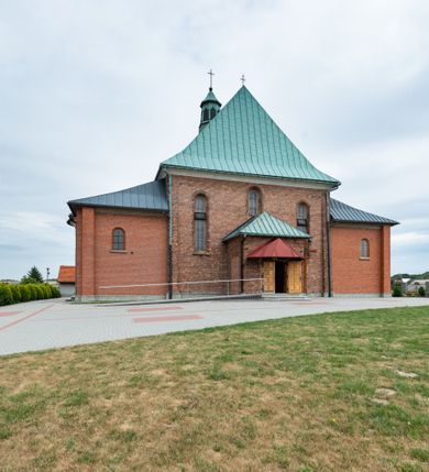 Zdjęcie nr 39: Kościół murowany z cegły na niskiej, kamiennej podmurówce, orientowany, jednonawowy ze zredukowanym transeptem oraz nieco niższym, zamkniętym półkoliście prezbiterium. Do północno-wschodniego narożnika nawy (przy prezbiterium) przylega założona na rzucie prostokąta, oszkarpowana zakrystia dostępna półkolistym portalem od strony północnej. Do południowo-wschodniego narożnika nawy dostawiona kaplica Matki Boskiej na rzucie prostokąta zamknięta od wschodu niewielką absydą. Ramiona transeptu zredukowane do roli prostokątnych kaplic. Do południowo-zachodniego i północno-zachodniego narożnika nawy dobudowane nowsze kruchty boczne założone na rzucie prostokąta dostępne portalami o półkolistym wykroju. Kruchta zachodnia na rzucie prostokąta dostępna nowym, prostokątnym wejściem. W ścianach północnej i południowej widoczne, zamurowane, starsze otwory wejściowe (od południa w miejsce portalu okrągłe okno). Dach nad nawą dwuspadowy z trzecią połacią zakrywającą ścianę szczytową; w części wschodniej wieloboczna wieżyczka na sygnaturkę z glorietą zwieńczoną krzyżem. Kruchty oraz kaplice boczne kryte dachami trójpołaciowymi, prezbiterium jednopołaciowym; nad zakrystią dach mansardowy z prostokątnym oknem w lukarnie. Pokrycia dachowe wykonane są z blachy (pierwotnie była to czerwona dachówka). W nawie i prezbiterium strop o konstrukcji żelbetowej wsparty na dziewięciu masywnych kolumnach ustawionych na wysokich cokołach; kaplice boczne sklepione krzyżowo, w zakrystii założony betonowy strop. Chór muzyczny murowany, wsparty na pierwszej od zachodu parze kolumn dźwigających strop. Fasada jednokondygnacyjna, trójosiowa z umieszczoną w osi centralnej prostopadłościenną kruchtą mieszczącą prostokątny otwór wejściowy (nowy). Okna w kształcie stojącego prostokąta o smukłych proporcjach, zamknięte półkoliście. Okno środkowe, umieszczone nieco wyżej, niższe od pozostałych. W ścianach zachodnich krucht bocznych po jednym niewielkim oknie o wykroju półkolistym. Trójkątna ściana szczytowa zasłonięta blaszanym dachem o przełamanej w dolnej części połaci, zwieńczona niewielkim, metalowym krzyżem. Elewacje boczne jednokondygnacyjne, o nieregularnym przebiegu osi. W elewacji południowej w pierwszej osi ceglany portal z nadświetlem o wykroju półkolistym, w drugiej okrągłe okno doświetlające kaplicę boczną, w kolejnych: półkolisty ceglany portal do zakrystii oraz okno o półkolistym wykroju z motywem biforium. W elewacji północnej w osi pierwszej i drugiej niewielkie półkoliście wykrojone okna, w osi trzeciej okrągłe okno doświetlające kaplicę, w osi czwartej natomiast ceglany portal  z nadświetlem o wykroju półkolistym. W prezbiterium cztery okna o wykroju półkolistym. W trzeciej osi, pomiędzy oknami, ujęta wielobocznymi filarami dźwigającymi, wydatny, profilowany gzyms tynkowana na biało nisza z figurą Matki Boskiej Fatimskiej umieszczonej pod baldachimem. Górną część murów nawy, prezbiterium, zakrystii, kaplic oraz obu krucht bocznych obiega profilowany gzyms okapowy. 