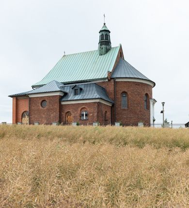 Zdjęcie nr 5: Kościół murowany z cegły na niskiej, kamiennej podmurówce, orientowany, jednonawowy ze zredukowanym transeptem oraz nieco niższym, zamkniętym półkoliście prezbiterium. Do północno-wschodniego narożnika nawy (przy prezbiterium) przylega założona na rzucie prostokąta, oszkarpowana zakrystia dostępna półkolistym portalem od strony północnej. Do południowo-wschodniego narożnika nawy dostawiona kaplica Matki Boskiej na rzucie prostokąta zamknięta od wschodu niewielką absydą. Ramiona transeptu zredukowane do roli prostokątnych kaplic. Do południowo-zachodniego i północno-zachodniego narożnika nawy dobudowane nowsze kruchty boczne założone na rzucie prostokąta dostępne portalami o półkolistym wykroju. Kruchta zachodnia na rzucie prostokąta dostępna nowym, prostokątnym wejściem. W ścianach północnej i południowej widoczne, zamurowane, starsze otwory wejściowe (od południa w miejsce portalu okrągłe okno). Dach nad nawą dwuspadowy z trzecią połacią zakrywającą ścianę szczytową; w części wschodniej wieloboczna wieżyczka na sygnaturkę z glorietą zwieńczoną krzyżem. Kruchty oraz kaplice boczne kryte dachami trójpołaciowymi, prezbiterium jednopołaciowym; nad zakrystią dach mansardowy z prostokątnym oknem w lukarnie. Pokrycia dachowe wykonane są z blachy (pierwotnie była to czerwona dachówka). W nawie i prezbiterium strop o konstrukcji żelbetowej wsparty na dziewięciu masywnych kolumnach ustawionych na wysokich cokołach; kaplice boczne sklepione krzyżowo, w zakrystii założony betonowy strop. Chór muzyczny murowany, wsparty na pierwszej od zachodu parze kolumn dźwigających strop. Fasada jednokondygnacyjna, trójosiowa z umieszczoną w osi centralnej prostopadłościenną kruchtą mieszczącą prostokątny otwór wejściowy (nowy). Okna w kształcie stojącego prostokąta o smukłych proporcjach, zamknięte półkoliście. Okno środkowe, umieszczone nieco wyżej, niższe od pozostałych. W ścianach zachodnich krucht bocznych po jednym niewielkim oknie o wykroju półkolistym. Trójkątna ściana szczytowa zasłonięta blaszanym dachem o przełamanej w dolnej części połaci, zwieńczona niewielkim, metalowym krzyżem. Elewacje boczne jednokondygnacyjne, o nieregularnym przebiegu osi. W elewacji południowej w pierwszej osi ceglany portal z nadświetlem o wykroju półkolistym, w drugiej okrągłe okno doświetlające kaplicę boczną, w kolejnych: półkolisty ceglany portal do zakrystii oraz okno o półkolistym wykroju z motywem biforium. W elewacji północnej w osi pierwszej i drugiej niewielkie półkoliście wykrojone okna, w osi trzeciej okrągłe okno doświetlające kaplicę, w osi czwartej natomiast ceglany portal  z nadświetlem o wykroju półkolistym. W prezbiterium cztery okna o wykroju półkolistym. W trzeciej osi, pomiędzy oknami, ujęta wielobocznymi filarami dźwigającymi, wydatny, profilowany gzyms tynkowana na biało nisza z figurą Matki Boskiej Fatimskiej umieszczonej pod baldachimem. Górną część murów nawy, prezbiterium, zakrystii, kaplic oraz obu krucht bocznych obiega profilowany gzyms okapowy. 