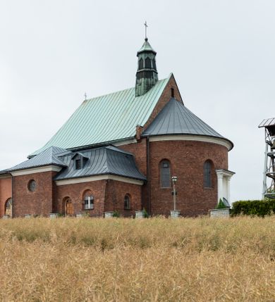 Zdjęcie nr 6: Kościół murowany z cegły na niskiej, kamiennej podmurówce, orientowany, jednonawowy ze zredukowanym transeptem oraz nieco niższym, zamkniętym półkoliście prezbiterium. Do północno-wschodniego narożnika nawy (przy prezbiterium) przylega założona na rzucie prostokąta, oszkarpowana zakrystia dostępna półkolistym portalem od strony północnej. Do południowo-wschodniego narożnika nawy dostawiona kaplica Matki Boskiej na rzucie prostokąta zamknięta od wschodu niewielką absydą. Ramiona transeptu zredukowane do roli prostokątnych kaplic. Do południowo-zachodniego i północno-zachodniego narożnika nawy dobudowane nowsze kruchty boczne założone na rzucie prostokąta dostępne portalami o półkolistym wykroju. Kruchta zachodnia na rzucie prostokąta dostępna nowym, prostokątnym wejściem. W ścianach północnej i południowej widoczne, zamurowane, starsze otwory wejściowe (od południa w miejsce portalu okrągłe okno). Dach nad nawą dwuspadowy z trzecią połacią zakrywającą ścianę szczytową; w części wschodniej wieloboczna wieżyczka na sygnaturkę z glorietą zwieńczoną krzyżem. Kruchty oraz kaplice boczne kryte dachami trójpołaciowymi, prezbiterium jednopołaciowym; nad zakrystią dach mansardowy z prostokątnym oknem w lukarnie. Pokrycia dachowe wykonane są z blachy (pierwotnie była to czerwona dachówka). W nawie i prezbiterium strop o konstrukcji żelbetowej wsparty na dziewięciu masywnych kolumnach ustawionych na wysokich cokołach; kaplice boczne sklepione krzyżowo, w zakrystii założony betonowy strop. Chór muzyczny murowany, wsparty na pierwszej od zachodu parze kolumn dźwigających strop. Fasada jednokondygnacyjna, trójosiowa z umieszczoną w osi centralnej prostopadłościenną kruchtą mieszczącą prostokątny otwór wejściowy (nowy). Okna w kształcie stojącego prostokąta o smukłych proporcjach, zamknięte półkoliście. Okno środkowe, umieszczone nieco wyżej, niższe od pozostałych. W ścianach zachodnich krucht bocznych po jednym niewielkim oknie o wykroju półkolistym. Trójkątna ściana szczytowa zasłonięta blaszanym dachem o przełamanej w dolnej części połaci, zwieńczona niewielkim, metalowym krzyżem. Elewacje boczne jednokondygnacyjne, o nieregularnym przebiegu osi. W elewacji południowej w pierwszej osi ceglany portal z nadświetlem o wykroju półkolistym, w drugiej okrągłe okno doświetlające kaplicę boczną, w kolejnych: półkolisty ceglany portal do zakrystii oraz okno o półkolistym wykroju z motywem biforium. W elewacji północnej w osi pierwszej i drugiej niewielkie półkoliście wykrojone okna, w osi trzeciej okrągłe okno doświetlające kaplicę, w osi czwartej natomiast ceglany portal  z nadświetlem o wykroju półkolistym. W prezbiterium cztery okna o wykroju półkolistym. W trzeciej osi, pomiędzy oknami, ujęta wielobocznymi filarami dźwigającymi, wydatny, profilowany gzyms tynkowana na biało nisza z figurą Matki Boskiej Fatimskiej umieszczonej pod baldachimem. Górną część murów nawy, prezbiterium, zakrystii, kaplic oraz obu krucht bocznych obiega profilowany gzyms okapowy. 