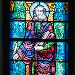 Zdjęcie nr 1: Witraż w kształcie stojącego prostokąta zamkniętego u góry łukiem półokrągłym. Kompozycja dwustrefowa. W dolnej części przedstawiony św. Łukasz ukazany frontalnie w ujęciu całopostaciowym. Święty wyobrażony jako młody mężczyzna o dużych, ciemnych oczach i długim, prostym nosie. Włosy sięgające ramion i krótka broda skręcone w spiralne loki. Postać ubrana w długą, zieloną tunikę oraz płaszcz w odcieniach brązu i fioletu trzymająca w prawej ręce pióro, w lewej zwój pergaminu. Spod szat widoczne bose stopy. W dolnej części kompozycji od lewej znajduje się wół – symbol tego ewangelisty oraz banderola z napisem: „ŚW(IĘTY) ŁUKASZ EWANGELISTA”. W tle widoczne wąskie romańskie kolumny o roślinnych kapitelach i trzonach dekorowanych geometryczną plecionką. W prześwitach pomiędzy kolumnami wkomponowane mniejsze sceny ukazujące wydarzenia z życia Marii i Chrystusa, opisane przez św. Łukasza. W górnej części kompozycji w arkadzie ujętej dwiema zdwojonymi kolumienkami ukazana Maria przedstawiona w pozie orantki. Postać ukazana frontalnie, w ujęciu całopostaciowym, ubrana w jasnoróżową suknię oraz błękitny płaszcz. Na głowie otoczonej nimbem biała chusta. Arkada, w której umieszczono Marię, ujęta jest motywami roślinnymi oraz dwiema mniejszymi arkadkami mieszczącymi motyw chrismon oraz monogram w postaci litery „M” zwieńczonej krzyżem.