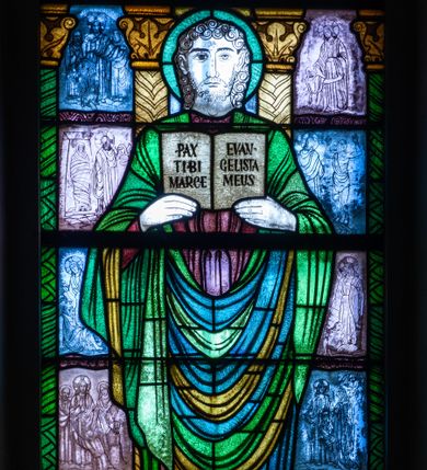 Zdjęcie nr 1: Witraż w kształcie stojącego prostokąta zamkniętego u góry łukiem półokrągłym. Kompozycja dwustrefowa. W dolnej części przedstawiony św. Marek ukazany frontalnie w ujęciu całopostaciowym. Święty wyobrażony jako młody mężczyzna o dużych, ciemnych oczach i długim, prostym nosie. Włosy i krótka broda skręcone w spiralne loki. Postać ubrana w długą, brązowo-fioletową tunikę oraz płaszcz w odcieniach zieleni, trzymająca w obu rękach uniesionych na wysokość piersi otwartą księgę z napisem „PAX TIBI MARCE / EVANGELISTA MEUS”. Spod szat widoczne stopy obute w sandały. W dolnej części kompozycji u stóp św. Marka widoczny lew – symbol ewangelisty oraz banderola z napisem „EWANGELISTA ŚW(IĘTY) MAREK”. W tle znajdują się wąskie romańskie kolumny o roślinnych kapitelach i trzonach dekorowanych geometryczną plecionką. W prześwitach pomiędzy kolumnami wkomponowane mniejsze sceny ukazujące cuda związane z publiczną działalnością Chrystusa opisane przez św. Marka. W górnej części kompozycji w arkadzie ujętej dwiema zdwojonymi kolumienkami z napisem „TU ES PETRUS” w archiwolcie ukazany św. Piotr stojący na łodzi oznaczonej znakiem krzyża. Święty ukazany frontalnie, w ujęciu całopostaciowym. Ubrany w jasnozieloną tunikę oraz rozwiany fioletowy płaszcz. Piotr w swoim lewej ręce trzyma klucz, prawą wspiera na lasce zwieńczonej krzyżem. Twarz pełna, oczy szeroko otwarte skierowane na wprost. Długa, ciemna broda, nad czołem wyraźna łysina. Arkada, w której umieszczono św. Piotra, ujęta jest motywami roślinnymi oraz dwiema mniejszymi arkadkami mieszczącymi przekrzyżowaną literę „P” z prawej strony kompozycji i krzyż osadzony na literze  „U” z lewej strony.