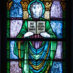 Zdjęcie nr 1: Witraż w kształcie stojącego prostokąta zamkniętego u góry łukiem półokrągłym. Kompozycja dwustrefowa. W dolnej części przedstawiony św. Marek ukazany frontalnie w ujęciu całopostaciowym. Święty wyobrażony jako młody mężczyzna o dużych, ciemnych oczach i długim, prostym nosie. Włosy i krótka broda skręcone w spiralne loki. Postać ubrana w długą, brązowo-fioletową tunikę oraz płaszcz w odcieniach zieleni, trzymająca w obu rękach uniesionych na wysokość piersi otwartą księgę z napisem „PAX TIBI MARCE / EVANGELISTA MEUS”. Spod szat widoczne stopy obute w sandały. W dolnej części kompozycji u stóp św. Marka widoczny lew – symbol ewangelisty oraz banderola z napisem „EWANGELISTA ŚW(IĘTY) MAREK”. W tle znajdują się wąskie romańskie kolumny o roślinnych kapitelach i trzonach dekorowanych geometryczną plecionką. W prześwitach pomiędzy kolumnami wkomponowane mniejsze sceny ukazujące cuda związane z publiczną działalnością Chrystusa opisane przez św. Marka. W górnej części kompozycji w arkadzie ujętej dwiema zdwojonymi kolumienkami z napisem „TU ES PETRUS” w archiwolcie ukazany św. Piotr stojący na łodzi oznaczonej znakiem krzyża. Święty ukazany frontalnie, w ujęciu całopostaciowym. Ubrany w jasnozieloną tunikę oraz rozwiany fioletowy płaszcz. Piotr w swoim lewej ręce trzyma klucz, prawą wspiera na lasce zwieńczonej krzyżem. Twarz pełna, oczy szeroko otwarte skierowane na wprost. Długa, ciemna broda, nad czołem wyraźna łysina. Arkada, w której umieszczono św. Piotra, ujęta jest motywami roślinnymi oraz dwiema mniejszymi arkadkami mieszczącymi przekrzyżowaną literę „P” z prawej strony kompozycji i krzyż osadzony na literze  „U” z lewej strony.