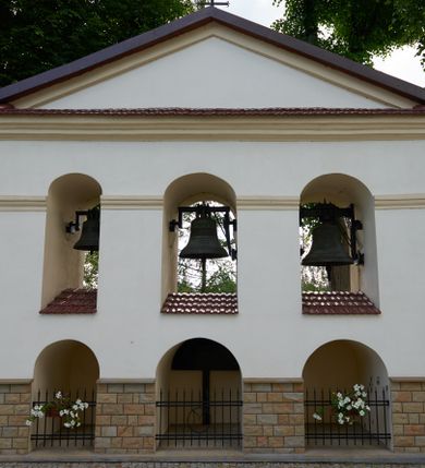 Zdjęcie nr 1: Dzwonnica typu parawanowego, wolnostojąca, murowana, tynkowana, trójosiowa. Obiekt wzniesiony na cokole obłożonym kamienną okładziną. W dolnej partii trzy arkady sięgające powyżej jej poziomu, oddzielone kratami. W górnej partii arkady przelotowe, od góry zamknięte w osi środkowej łukiem półkolistym, z przechodzącym w partii muru gzymsem. W każdej arkadzie górnej wisi jeden dzwon; arkady obu kondygnacji lewej osi mniejsze. Dzwonnicę wieńczy gzyms podokapowy, powyżej którego trójkątny szczyt podkreślony dekoracją ramową i przekryty blachą.  