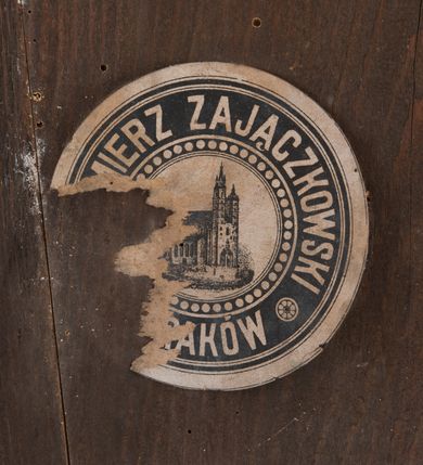 Zdjęcie nr 2: Zegar w kształcie stojącego prostokąta. W części centralnej okrągła tarcza, pośrodku kielich z hostią na tle błękitnego nieba, od którego odchodzą dwie fantazyjnie wycięte wskazówki. Wokół tarczy dwa okręgi, w pierwszym nazwy miesiąców, w drugim cyfry od 1 do 31. Powyżej napis „RENOVATIO * SANCTISSIMI * SACRAMENTI”, a nad nim dwa anioły podtrzymujące zamkniętą koronę. Anioły o owalnych twarzach, młodzieńczych rysach i średniej długości bujnych włosach. Ubrane w długie suknie z długimi rękawami, przewiązane w pasie wstążką. Rysunek oddany sumarycznie. W dolnej części kompozycji stylizowany, ornamentalny kartusz, w polu słaboczytelny napis i data „1906”. Poniżej sygnatury: „Nakładem K(azimierza) Zajączkowskiego w Krakowie.” oraz „Lit(itografia) A(?) S(D?)urczykiewicza w Krakowie.
