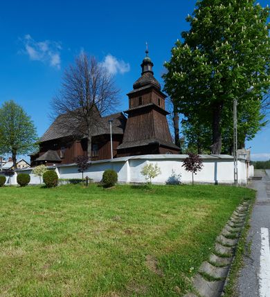 Zdjęcie nr 34: Kościół orientowany, drewniany, na podmurówce z kamienia łupanego, konstrukcji zrębowej, jednonawowy, szalowany, z wieżą konstrukcji słupowej usytuowaną od strony prezbiterium, z zakrystią w przyziemiu. Nawa prostokątna, szersza od prezbiterium, z kruchtami od zachodu, południa i północy. Prezbiterium zamknięte trójbocznie. Wieża od wschodu, na planie kwadratu, o konstrukcji ze słupów pochyłych, na zewnątrz trzy kondygnacje przedzielone obiegającymi daszkami. Dolna i górna izbica o ścianach pionowych, środkowa o ścianach pochyłych; wieża zwieńczona hełmem baniastym z latarnią. Kościół przykrywa dach siodłowy, na kruchtach trójspadowy, kryty cementówką. Pod wydatnymi okapami dachów biegnie gzyms o profilu ćwierćkoliście wklęsłym z wałkiem. Więźba dachowa nad prezbiterium jest krokwiowa, a nad nawą płatwiowa. Wewnątrz znajdują się stropy z fasetami, pod którymi umieszczony jest profilowany gzyms. Arkada tęczowa o łuku spłaszczonym. Chór muzyczny wsparty jest na dwóch filarach z wybrzuszoną balustradą. Wnętrze polichromowane, ściany w kruchtach oszalowane. Do kościoła prowadzą cztery wejścia: główne od zachodu, dwa boczne od południa i północy oraz przez zakrystię od wieży, od strony północno-zachodniej. W prezbiterium znajdują się dwa okna: po stronie południowej i północnej; a w nawie dwa po stronie południowej i północnej, natomiast w wieży cztery okna prostokątne, w latarni osiem okien zamkniętych dwoma nałęczami. Kościół otoczony jest murkiem ze stacjami drogi krzyżowej i dwiema bramkami z lat 1863 i 1864.