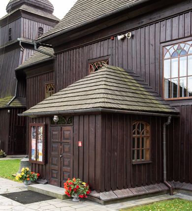 Zdjęcie nr 39: Kościół orientowany, drewniany, na podmurówce z kamienia łupanego, konstrukcji zrębowej, jednonawowy, szalowany, z wieżą konstrukcji słupowej usytuowaną od strony prezbiterium, z zakrystią w przyziemiu. Nawa prostokątna, szersza od prezbiterium, z kruchtami od zachodu, południa i północy. Prezbiterium zamknięte trójbocznie. Wieża od wschodu, na planie kwadratu, o konstrukcji ze słupów pochyłych, na zewnątrz trzy kondygnacje przedzielone obiegającymi daszkami. Dolna i górna izbica o ścianach pionowych, środkowa o ścianach pochyłych; wieża zwieńczona hełmem baniastym z latarnią. Kościół przykrywa dach siodłowy, na kruchtach trójspadowy, kryty cementówką. Pod wydatnymi okapami dachów biegnie gzyms o profilu ćwierćkoliście wklęsłym z wałkiem. Więźba dachowa nad prezbiterium jest krokwiowa, a nad nawą płatwiowa. Wewnątrz znajdują się stropy z fasetami, pod którymi umieszczony jest profilowany gzyms. Arkada tęczowa o łuku spłaszczonym. Chór muzyczny wsparty jest na dwóch filarach z wybrzuszoną balustradą. Wnętrze polichromowane, ściany w kruchtach oszalowane. Do kościoła prowadzą cztery wejścia: główne od zachodu, dwa boczne od południa i północy oraz przez zakrystię od wieży, od strony północno-zachodniej. W prezbiterium znajdują się dwa okna: po stronie południowej i północnej; a w nawie dwa po stronie południowej i północnej, natomiast w wieży cztery okna prostokątne, w latarni osiem okien zamkniętych dwoma nałęczami. Kościół otoczony jest murkiem ze stacjami drogi krzyżowej i dwiema bramkami z lat 1863 i 1864.